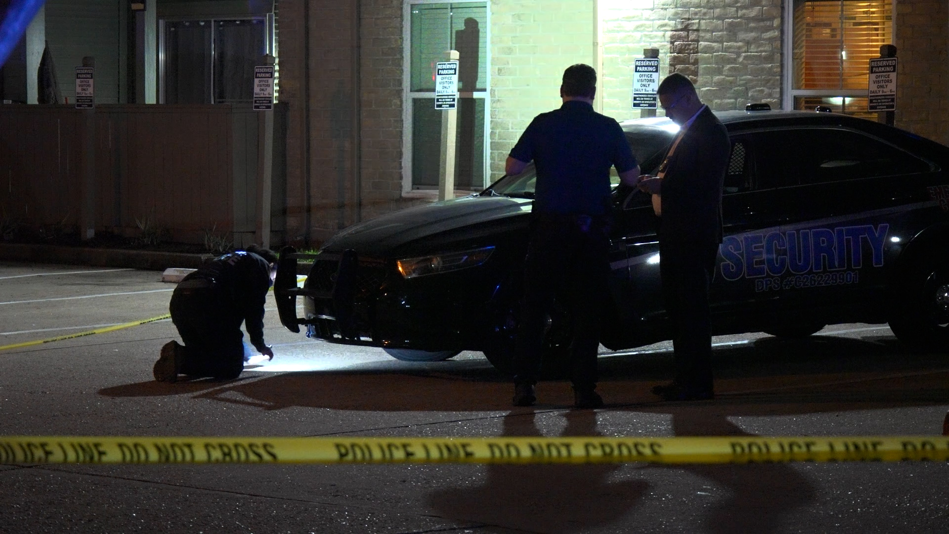 Nighttime crime scene with two officers near a black security patrol car, yellow tape in the foreground, and investigators examining the area.