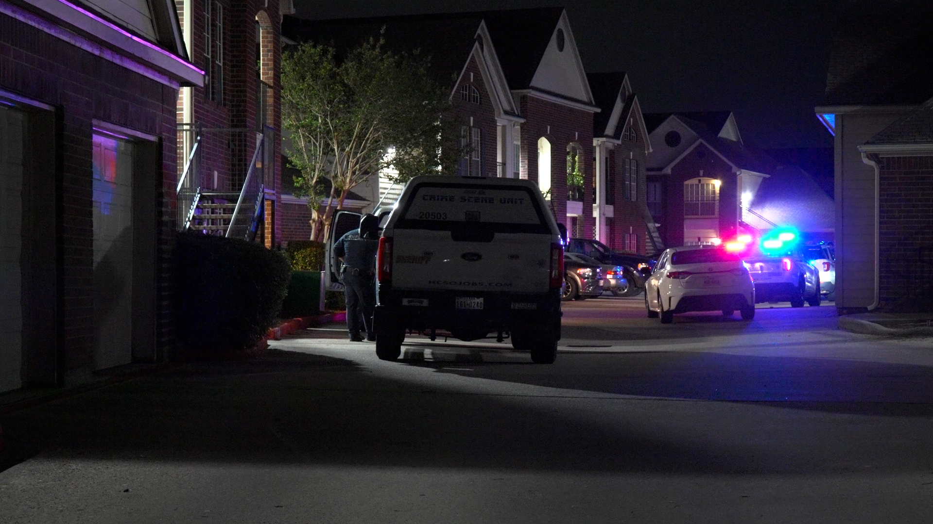Nighttime street scene with police cars flashing red and blue lights outside brick townhomes; a crime scene van is parked in the middle.