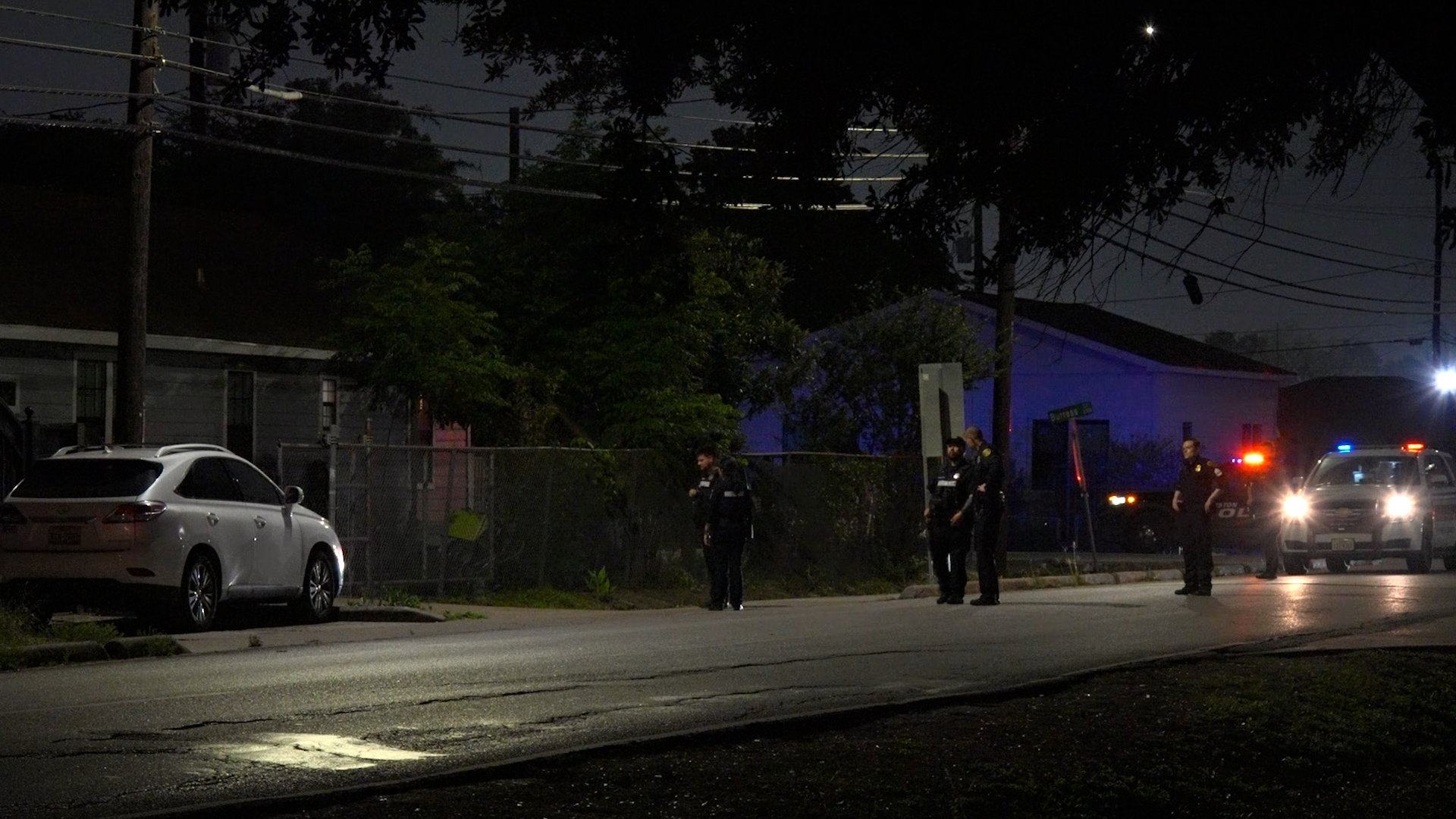 Nighttime street scene with police officers near a chain-link fence and a white sedan on the curb, police SUV lights flashing in the background.