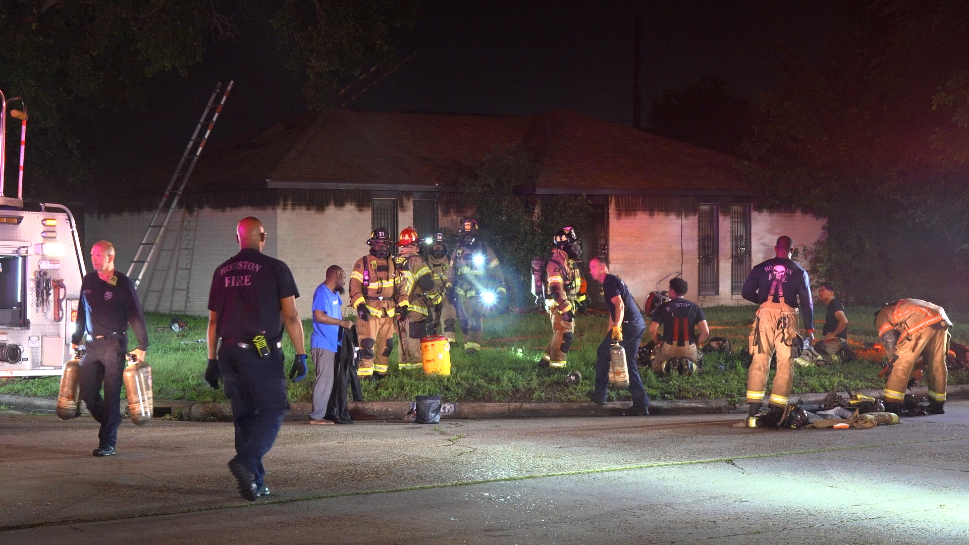 Nighttime fire scene with multiple firefighters in turnout gear and a fire truck on the left, ladder propped up, and responders handling equipment on a street.