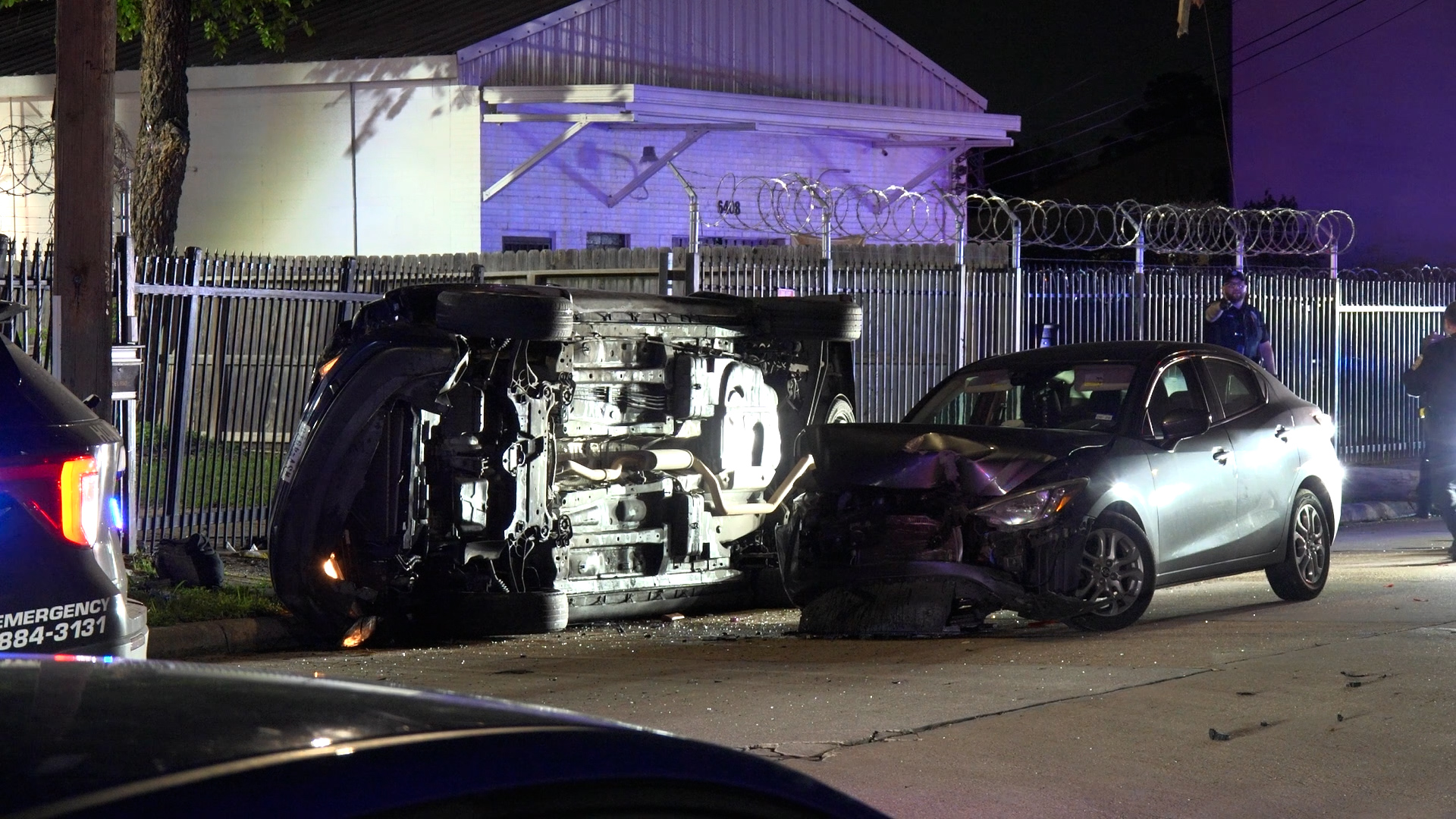 Nighttime car crash scene with a flipped SUV on its roof and a damaged silver sedan nearby, police officers in the background, barbed-wire fence, and emergency lights illuminating the area.