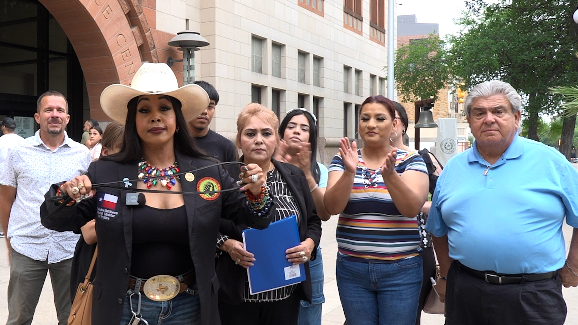 Group of diverse adults standing outside a brick building; a woman wearing a cowboy hat holds a lasso, while others applaud.