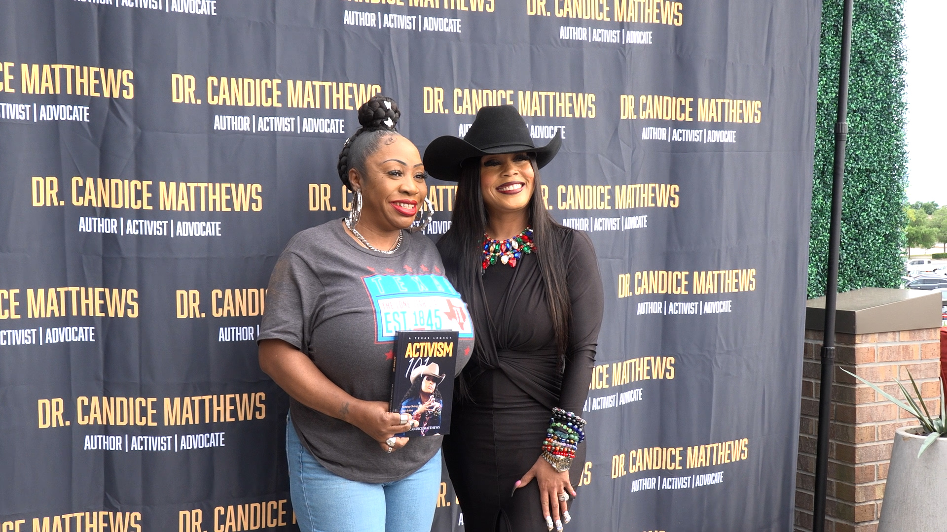 Two women pose at a Dr. Candice Matthews backdrop; the left woman in a gray shirt holds a book titled 'Activism', the right woman wears black with a hat and colorful jewelry.