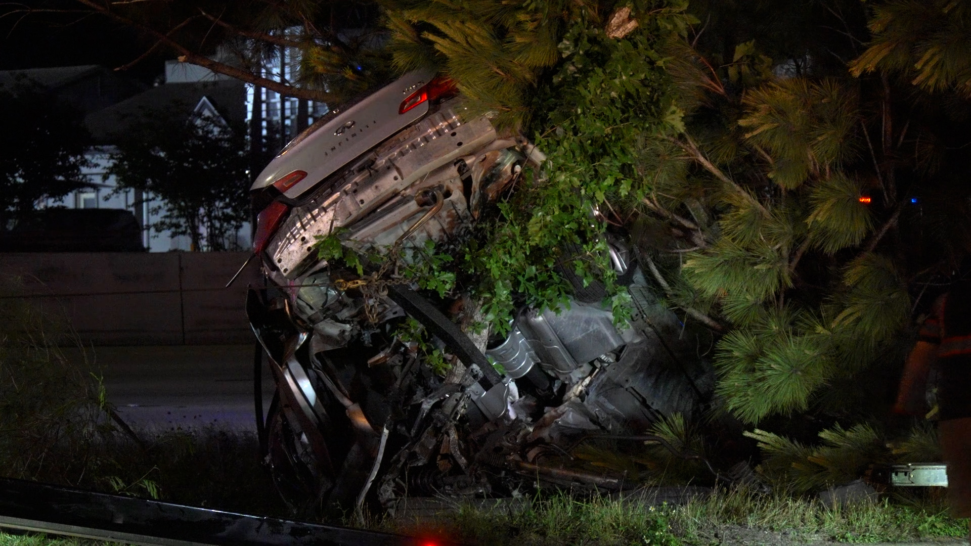 Silver sedan crushed against a large pine tree, wreckage strewn on the ground at night beside a roadway.
