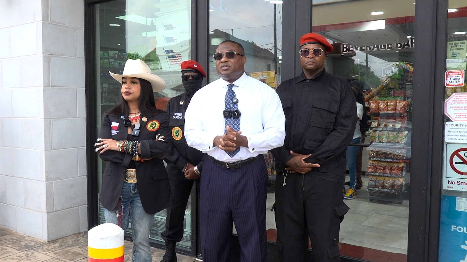 Four people stand outside a storefront; a man in a white shirt and patterned tie speaks while others listen.