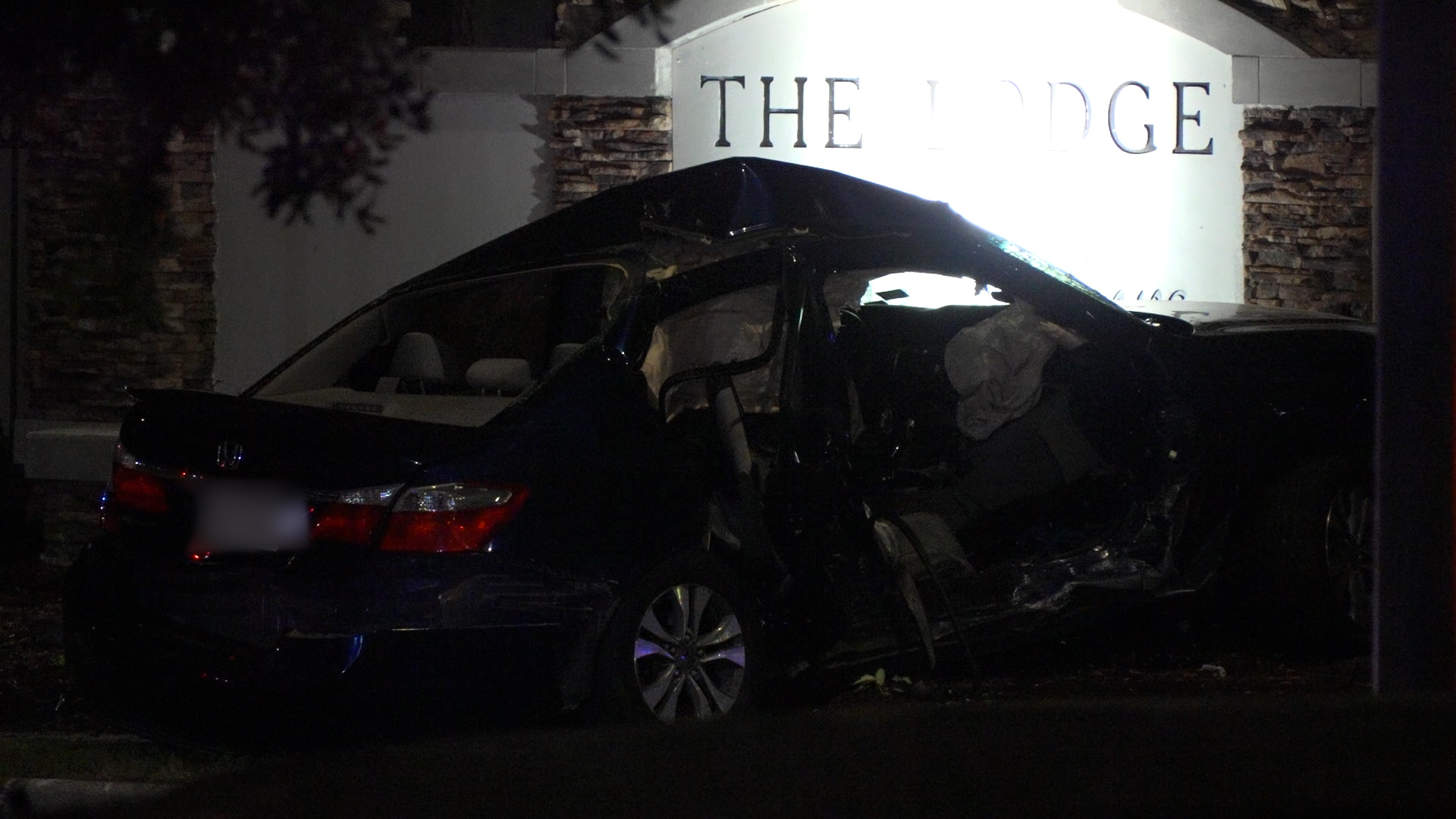 Nighttime scene of a severely damaged black car crashed into a stone wall and a lit wall sign that reads 'THE RIDGE' behind it; driver-side area is crushed and interior is exposed.