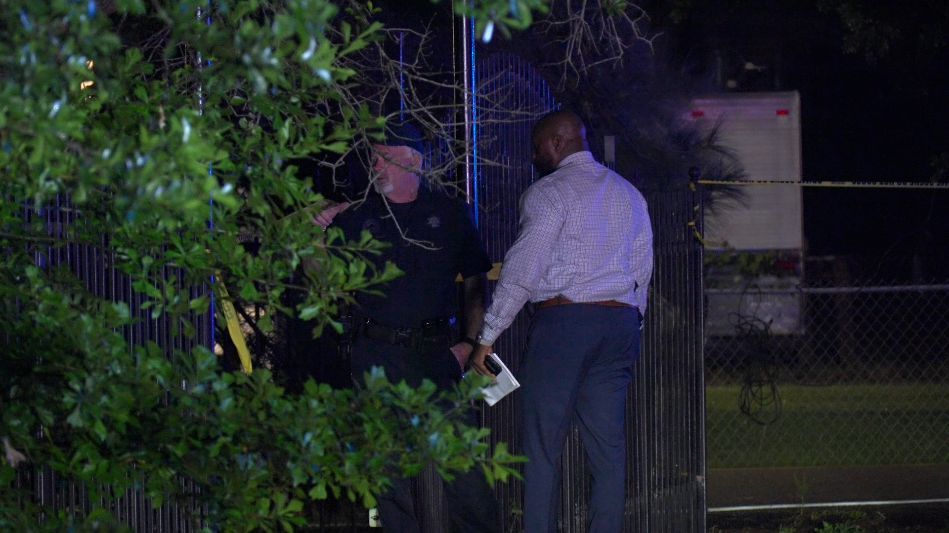 Two men speak by a tall metal gate at night, one in a police uniform and the other in a light shirt, holding papers; yellow caution tape in the background.