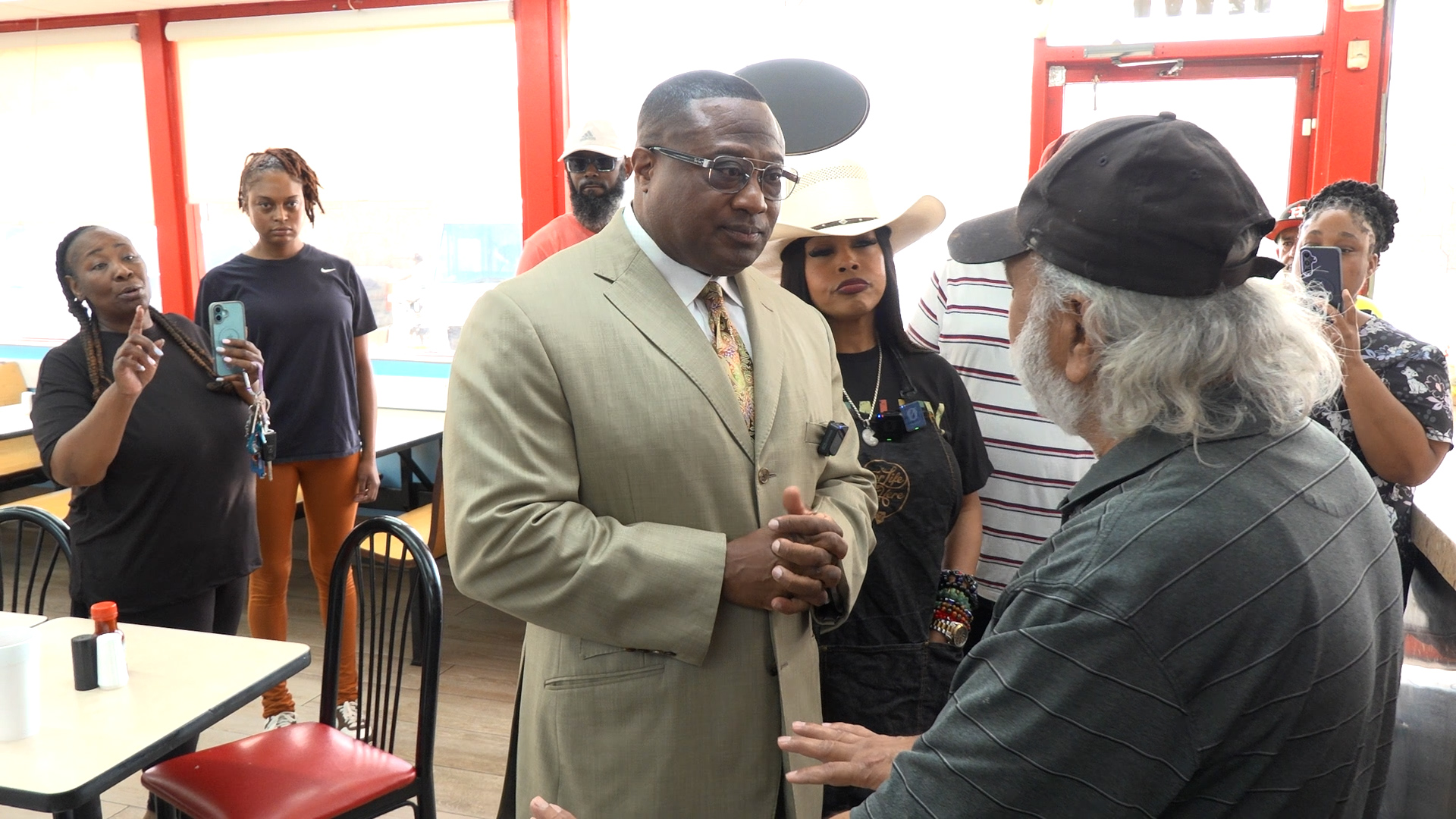 Man in a beige suit and glasses speaks to an older man wearing a cap, as a small crowd watches inside a diner-like space.
