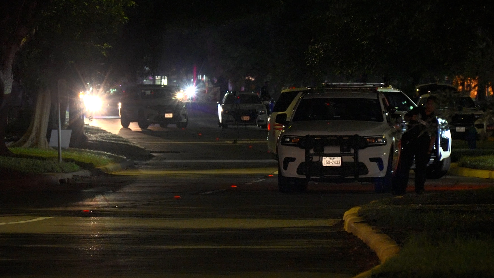 Nighttime street scene with police SUVs and flashing lights; an officer stands near a white SUV on a dimly lit road.