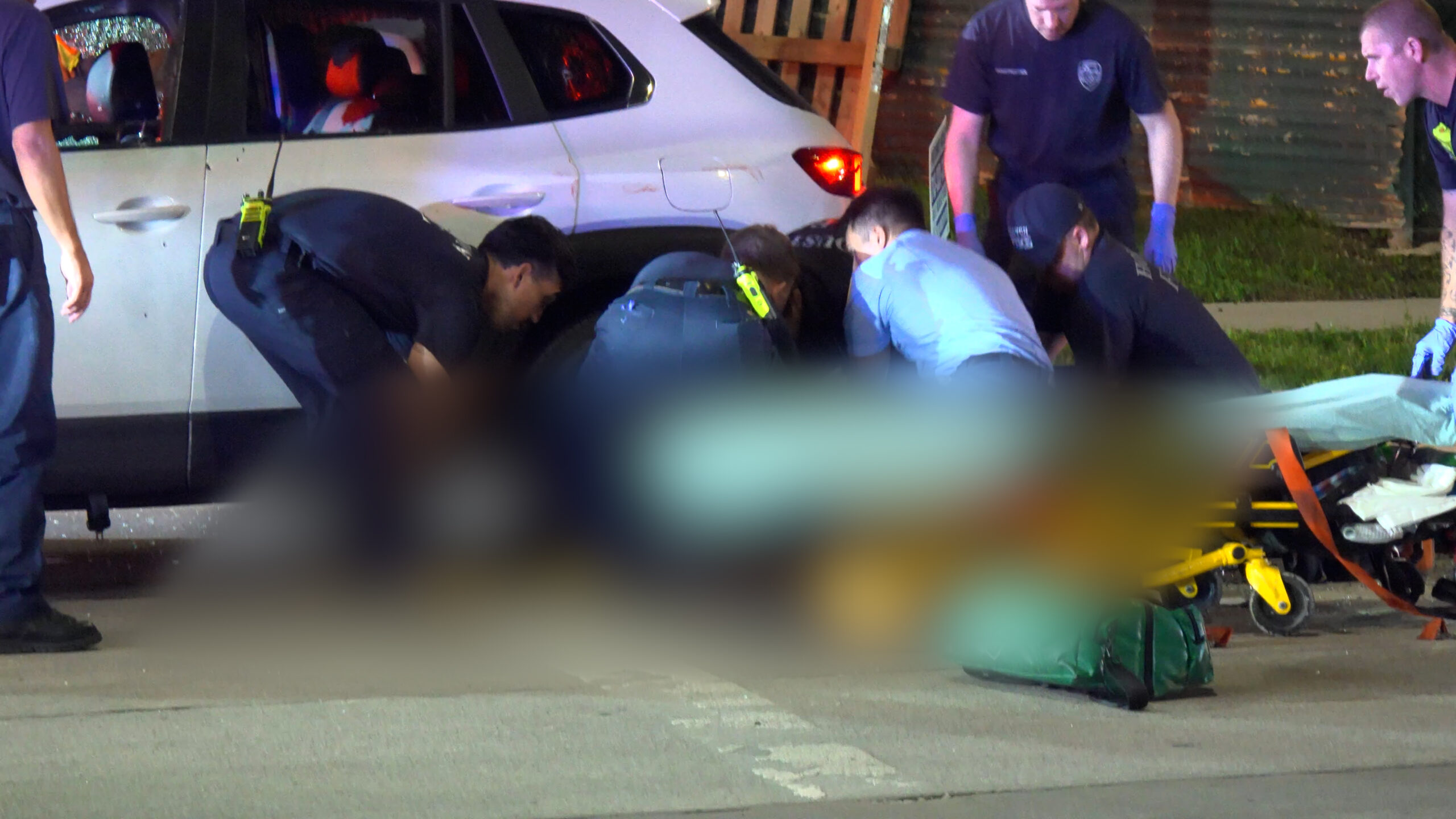 Emergency responders kneel by a white SUV at night, tending to a patient with a stretcher and medical gear on the ground.