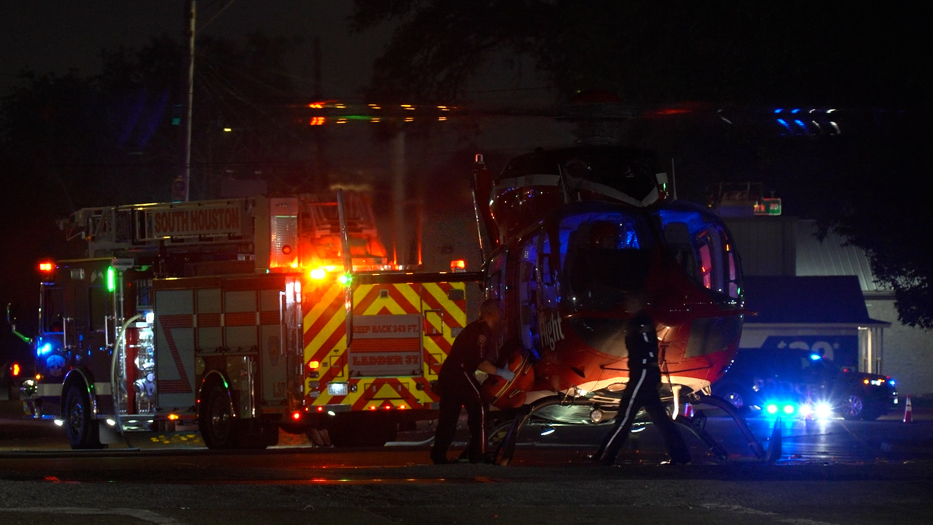 Night-time emergency scene with a red fire truck and a blue helicopter on the street, responders moving equipment under flashing lights.