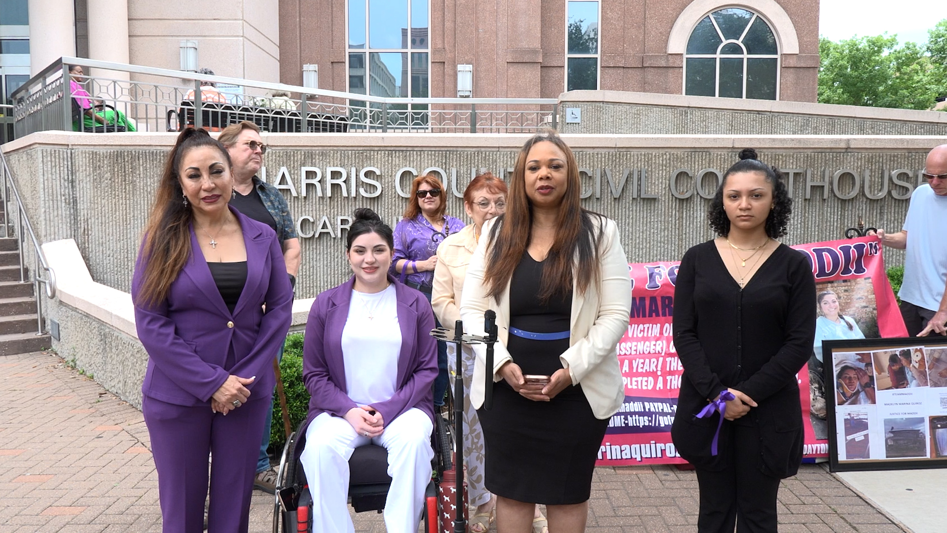 Group of women standing outside a courthouse, one at a microphone, two in purple suits, a woman in a wheelchair, with a protest banner in the background.