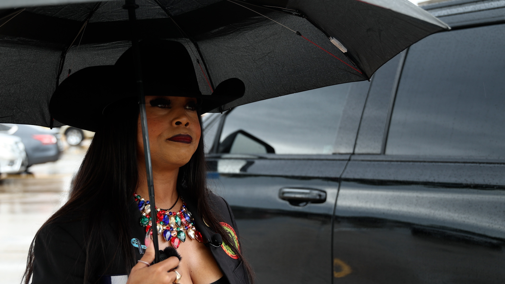 Woman with a black wide-brim hat and colorful beaded necklace holding a black umbrella in a rainy parking lot beside a dark vehicle, looking ahead.