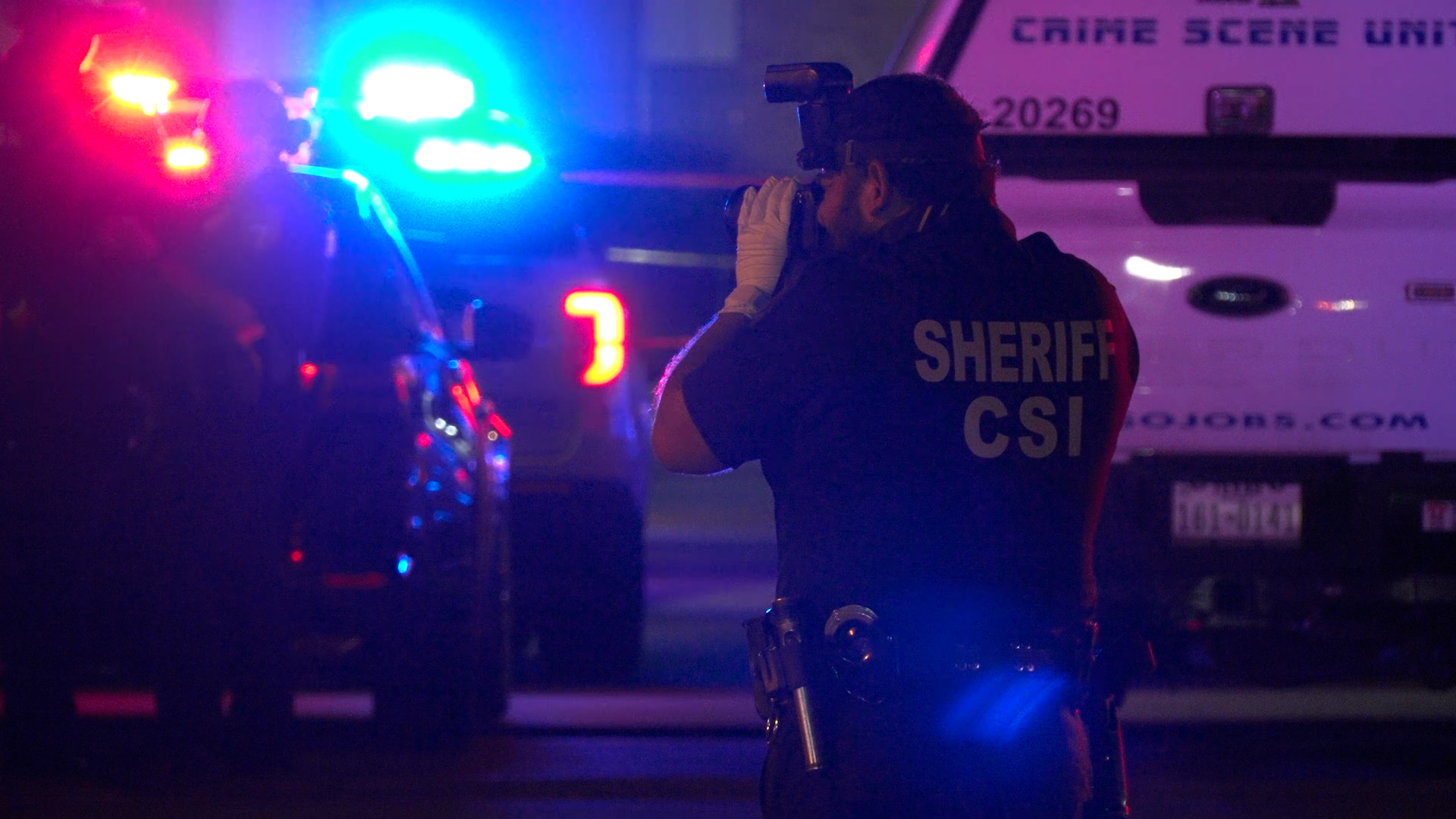Deputy from SHERIFF CSI photographing a crime scene at night, blue and red emergency lights flashing with a crime scene unit vehicle in the background.