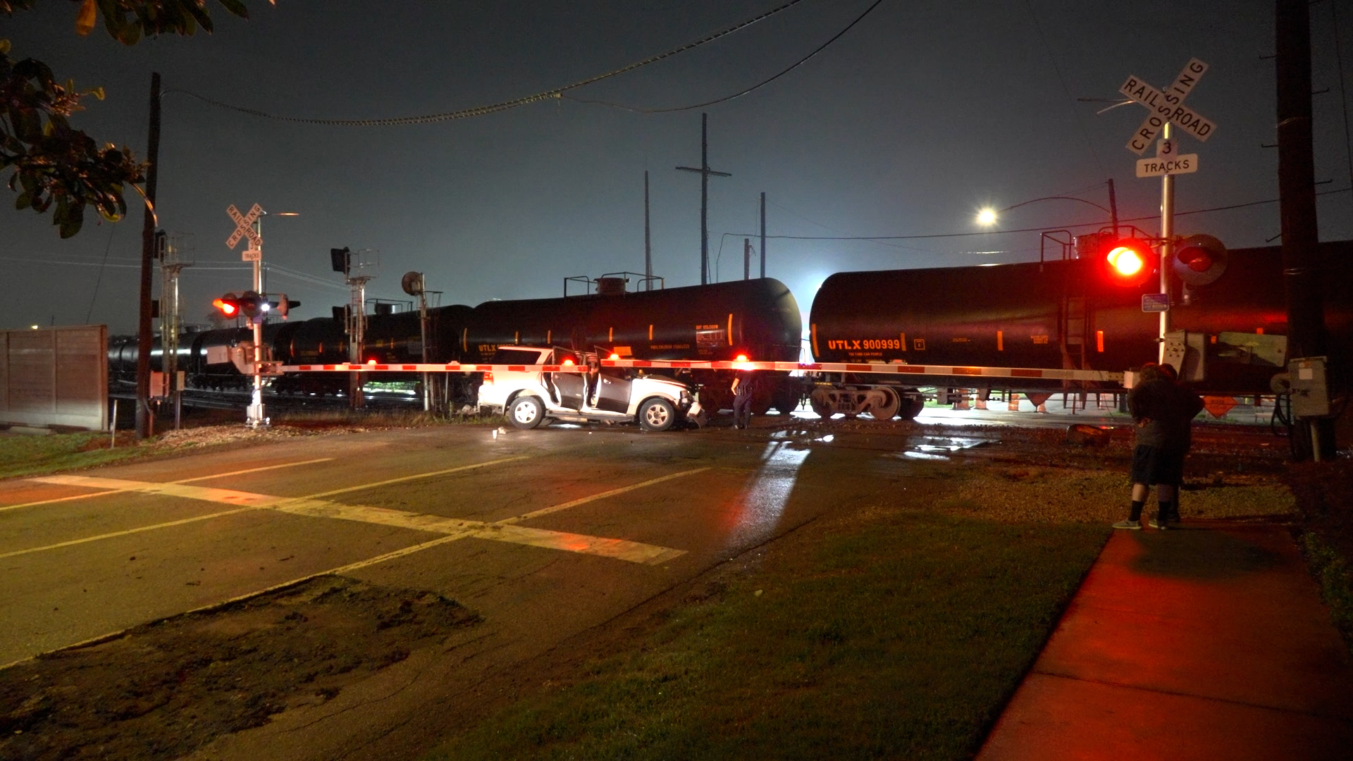 Nighttime crash at a railroad crossing: a white SUV is crushed near tanker cars with red emergency lights flashing in the scene's glow.
