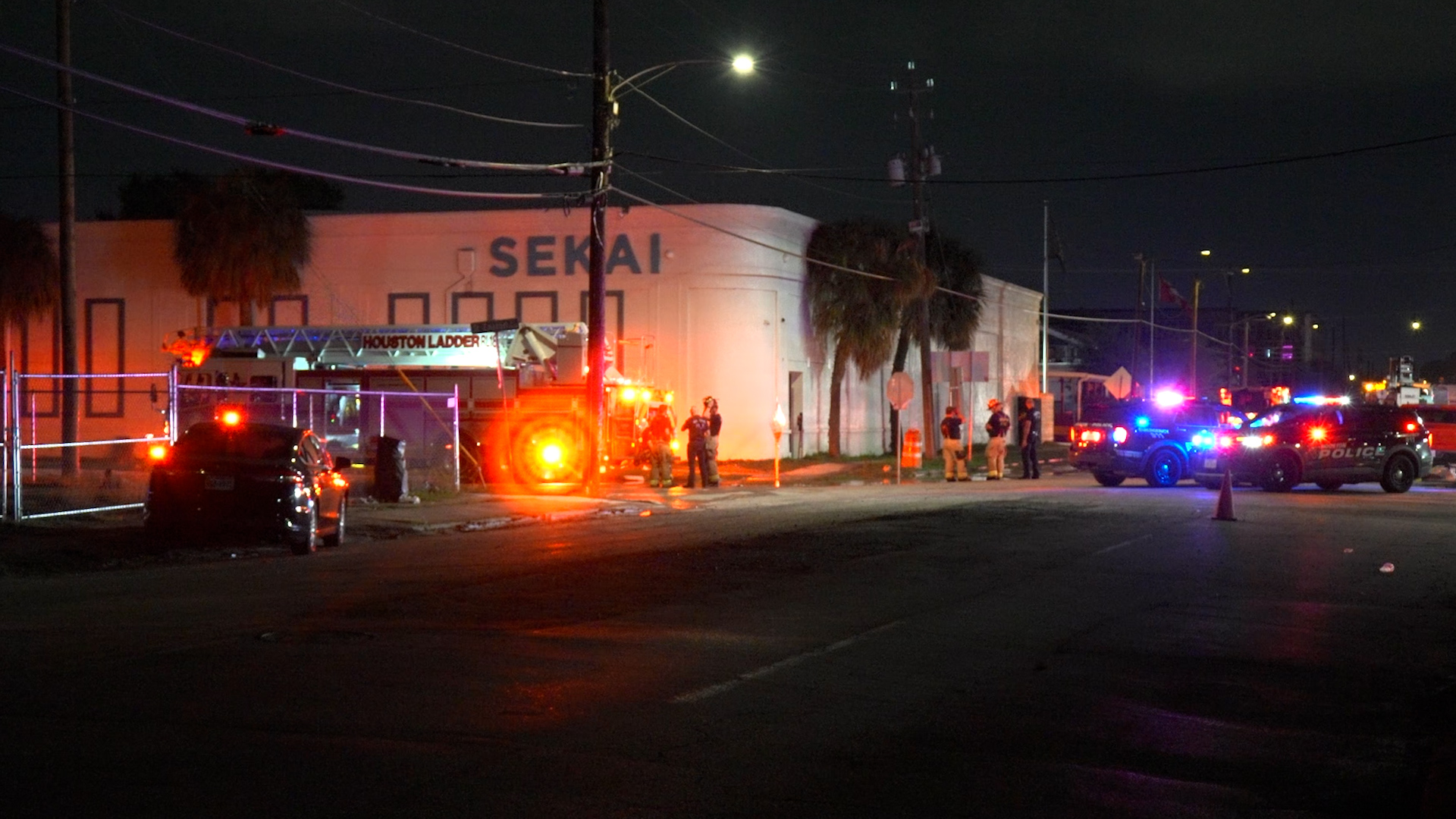 Nighttime street scene with a large building labeled 'SEKAI', emergency vehicles flashing red and blue lights, and responders nearby.