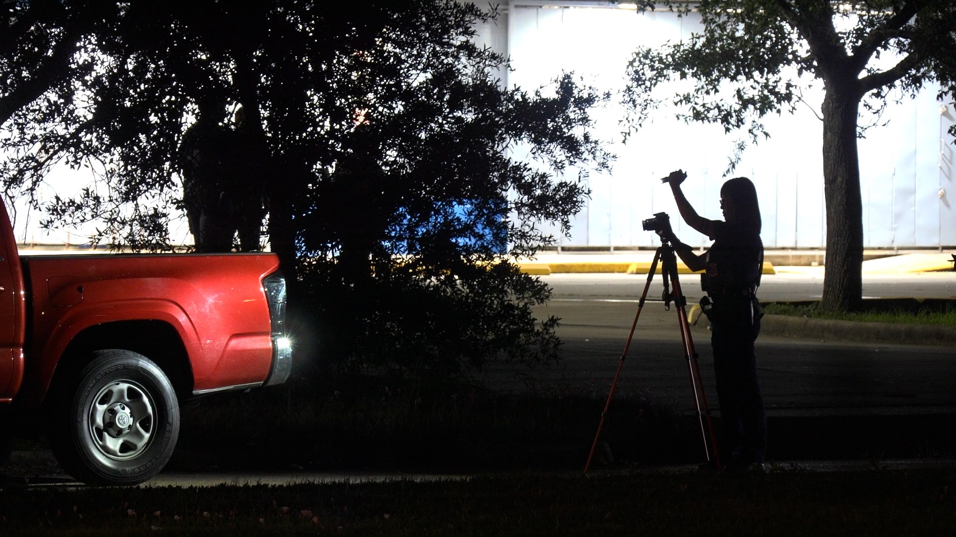 Photographer adjusting a camera on a tripod in a dim parking lot beside a red pickup truck.