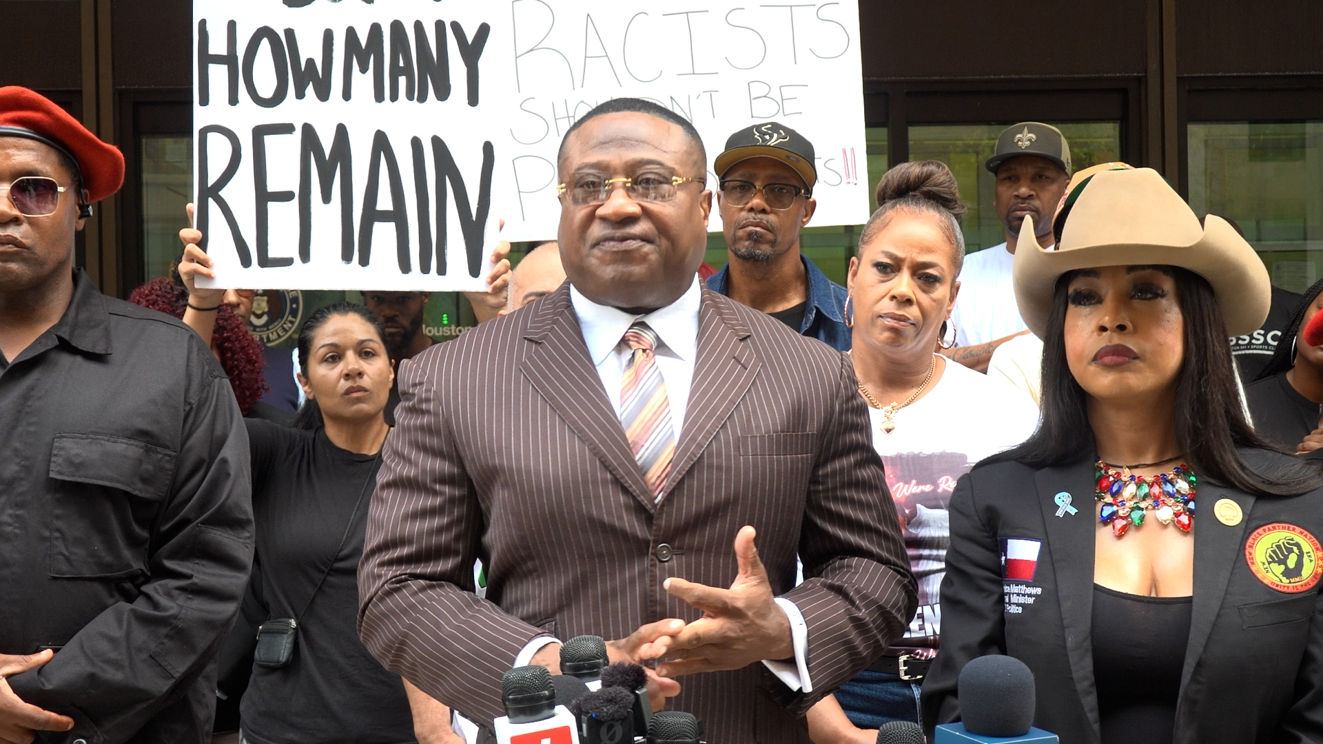 Group of protesters with a man in a pinstripe suit speaking at microphones, large sign behind reads 'HOW MANY REMAIN'.