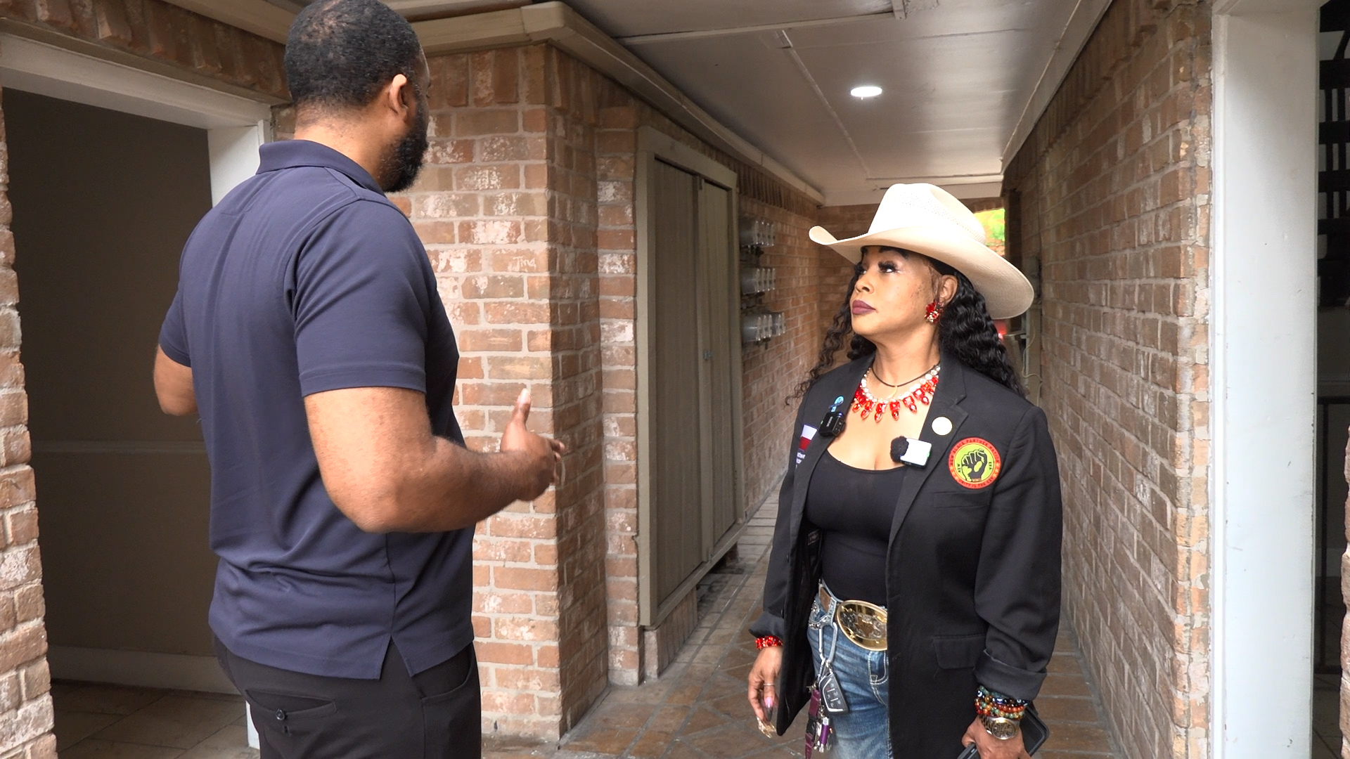 Man in a navy polo chats with a woman in a cream cowboy hat along a brick hallway.