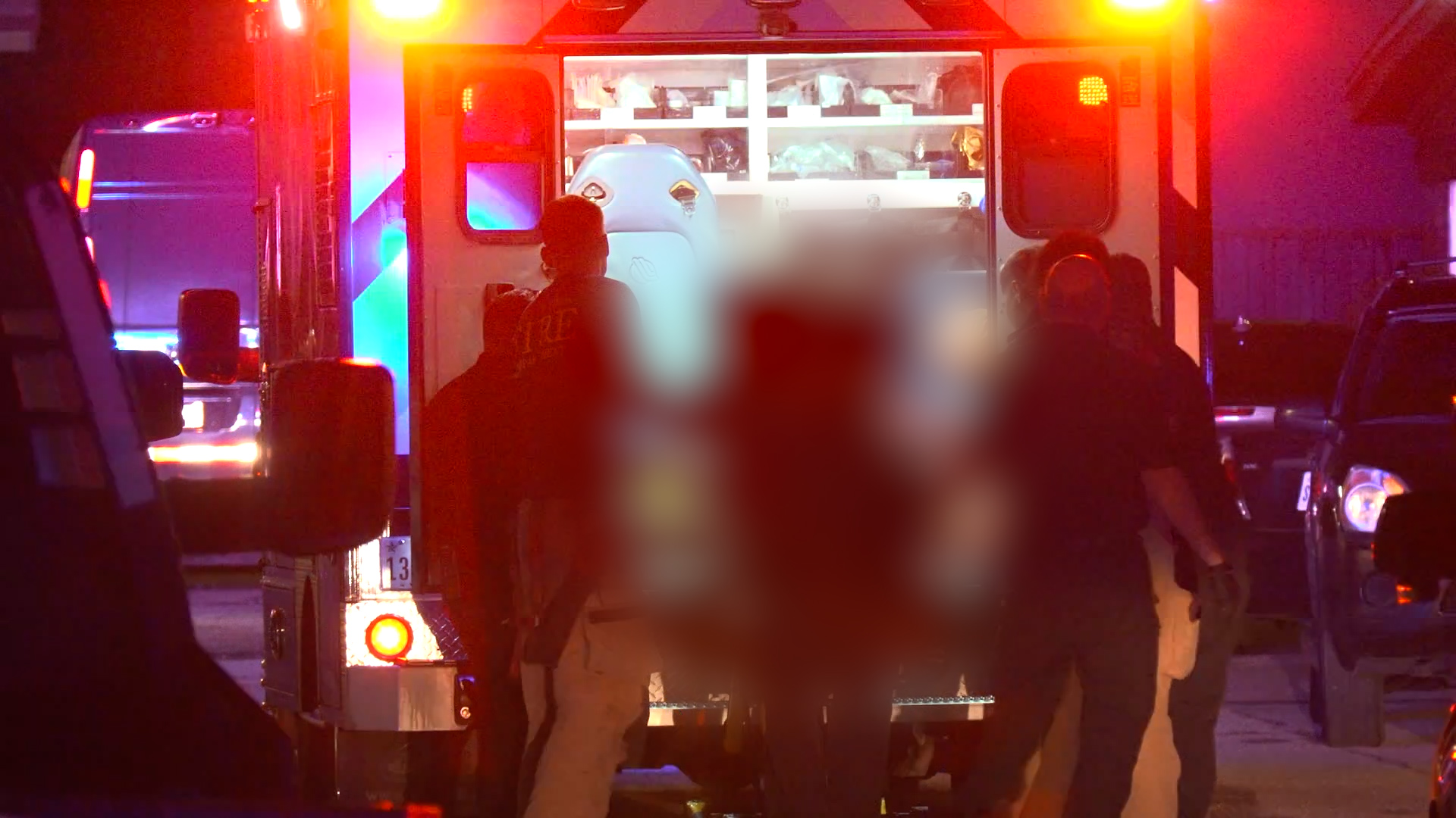 Nighttime emergency scene with responders by an ambulance, red and blue lights flashing as they attend to a patient area blurred for privacy.
