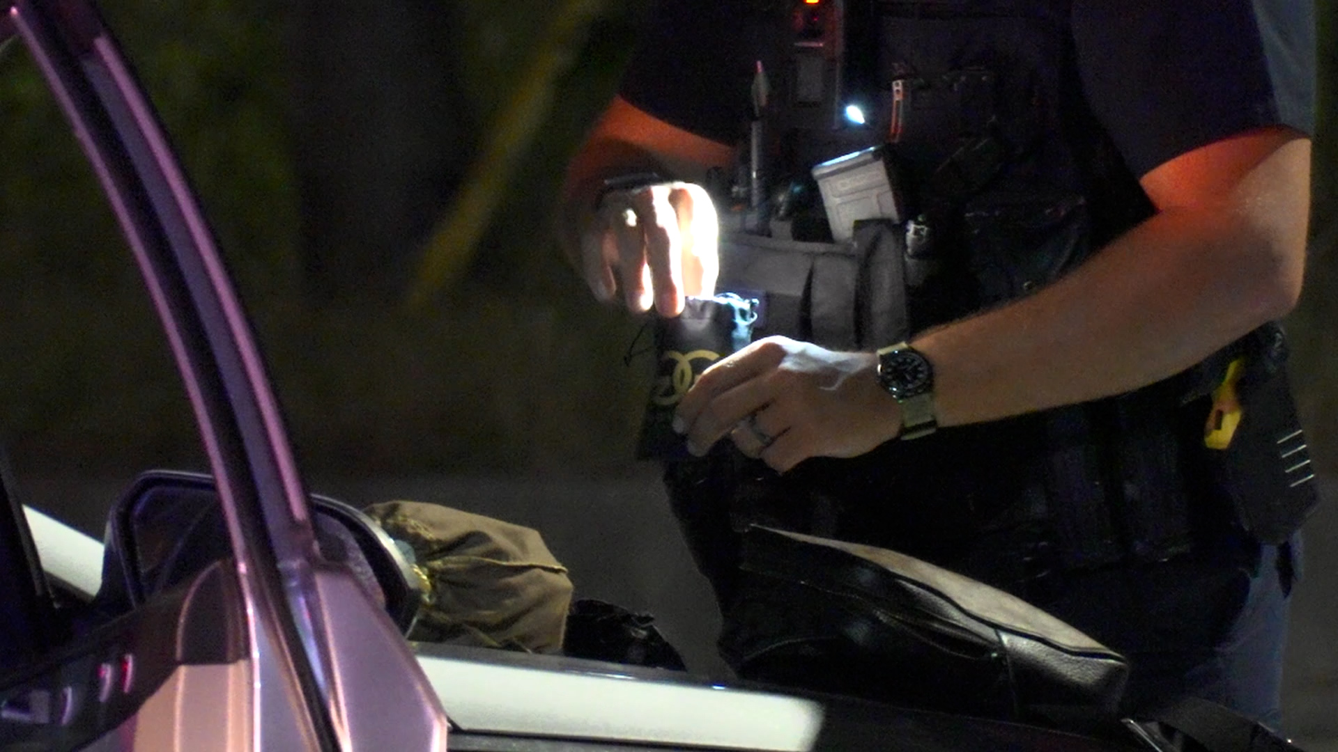 Police officer in tactical vest searches a small bag near a car window at night, hands lit by a flashlight or device light