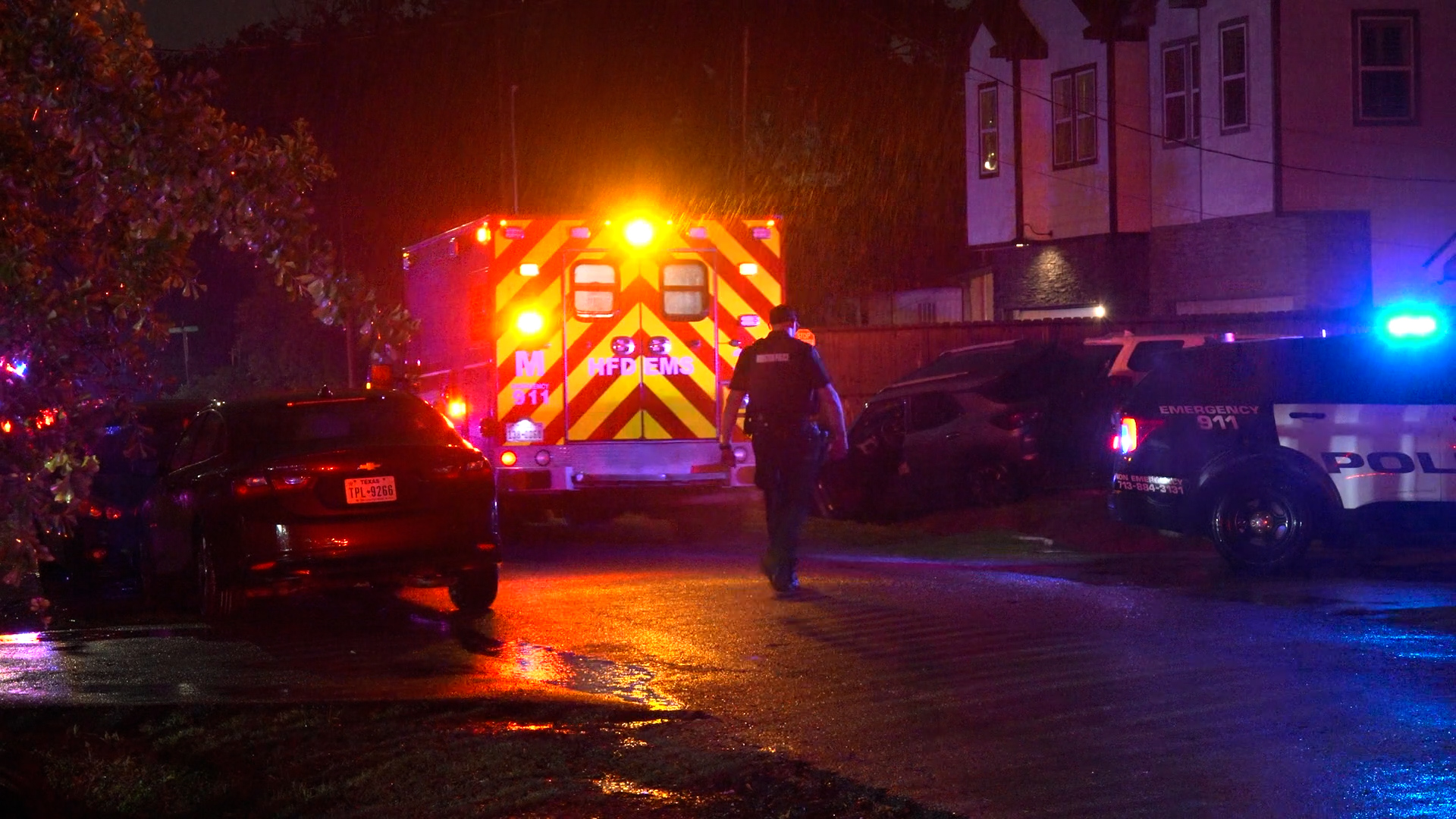Nighttime emergency scene with an ambulance's bright chevron back and flashing lights, a police SUV with blue lights, and an officer walking on a wet street.