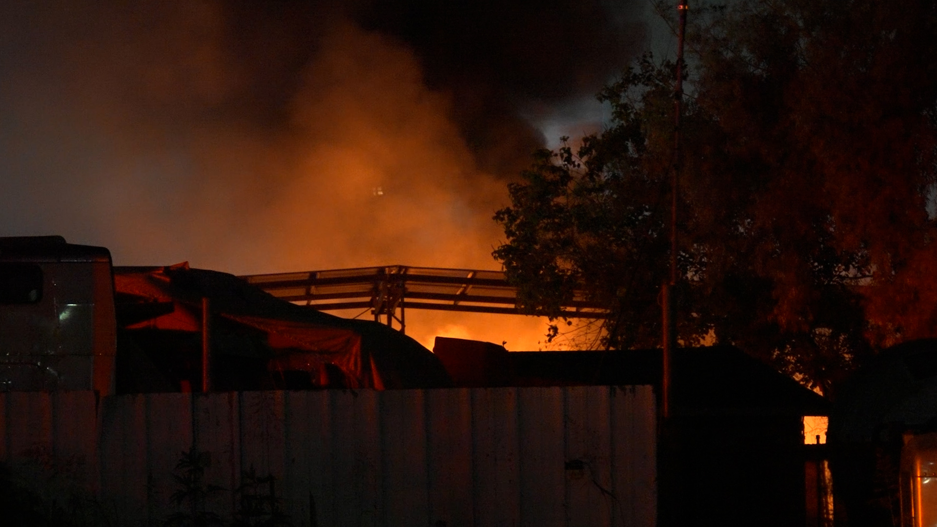 Nighttime scene of a structure on fire behind a wooden fence, with orange flames and dark smoke rising.