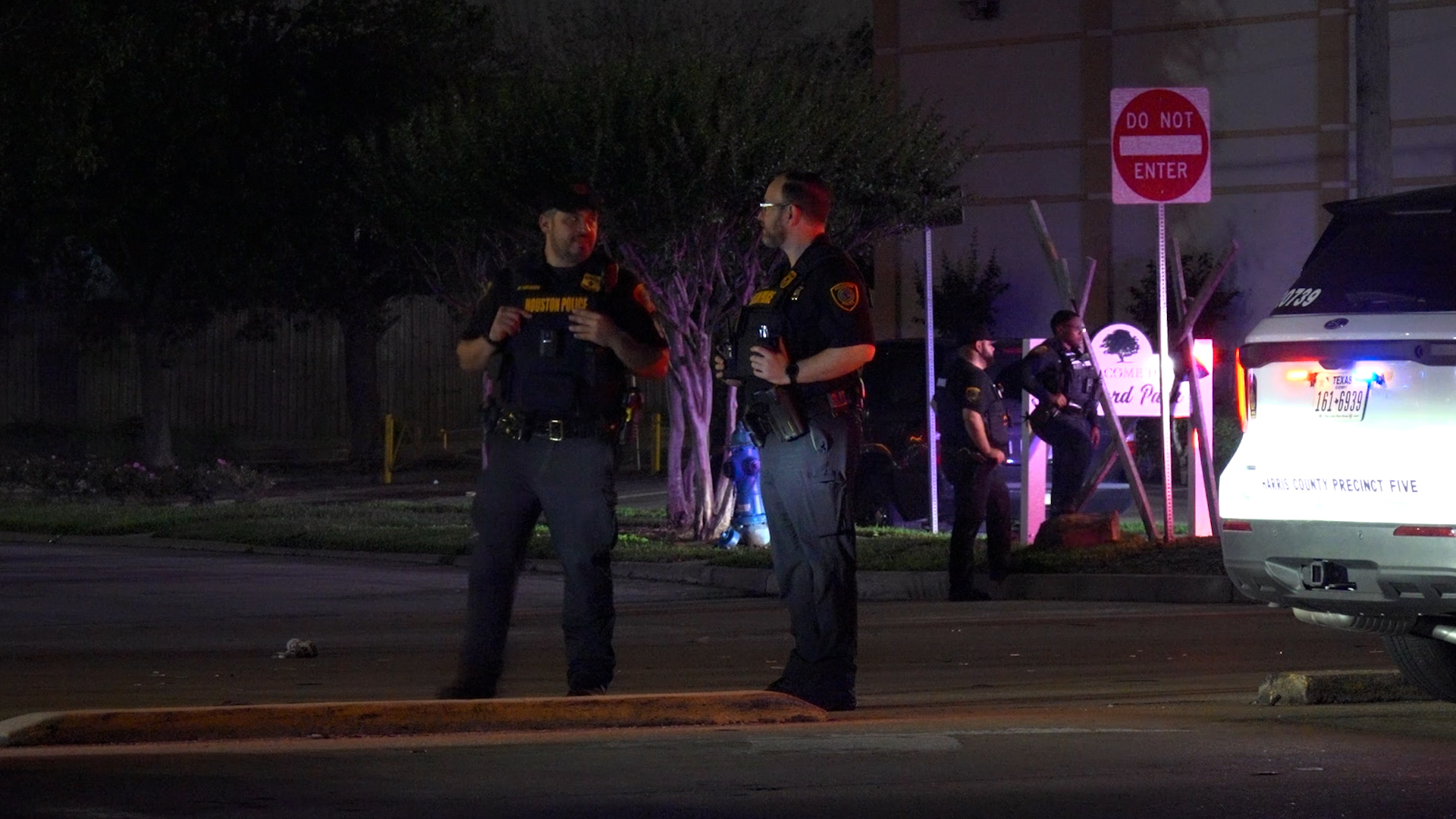 Nighttime scene with Houston police officers in tactical gear conversing near a marked police SUV on a dim street with red/blue lights nearby.