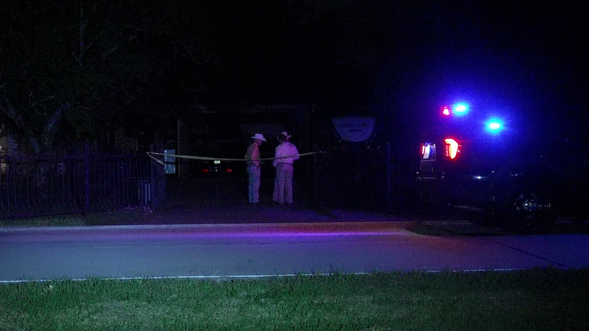 Nighttime street scene with two investigators standing behind police tape while a vehicle with flashing blue lights is nearby.