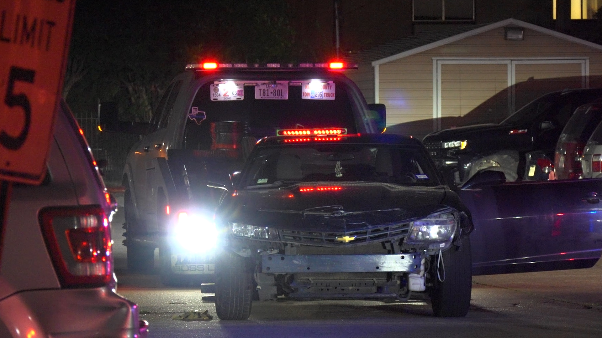 Nighttime multi-vehicle crash scene with a damaged black pickup in the foreground and red emergency lights flashing from a police or rescue vehicle in the background in a residential street setting.