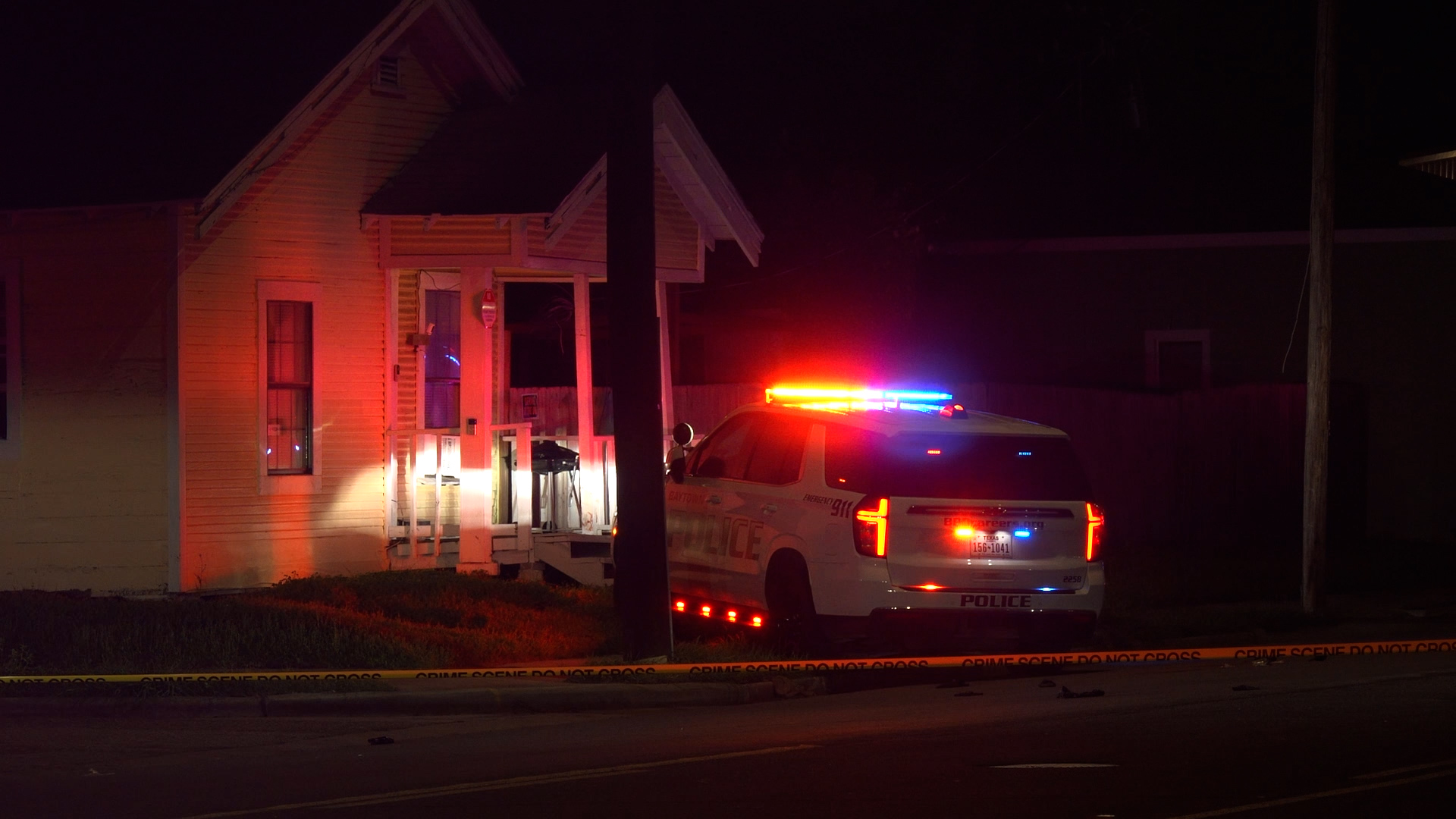 Police SUV with red and blue emergency lights at night in front of a house, crime scene tape across the street.