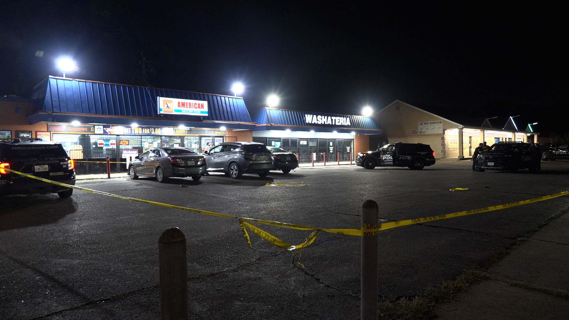 Nighttime parking lot of a strip mall with a blue-roofed store, police cars, and yellow caution tape across the foreground.