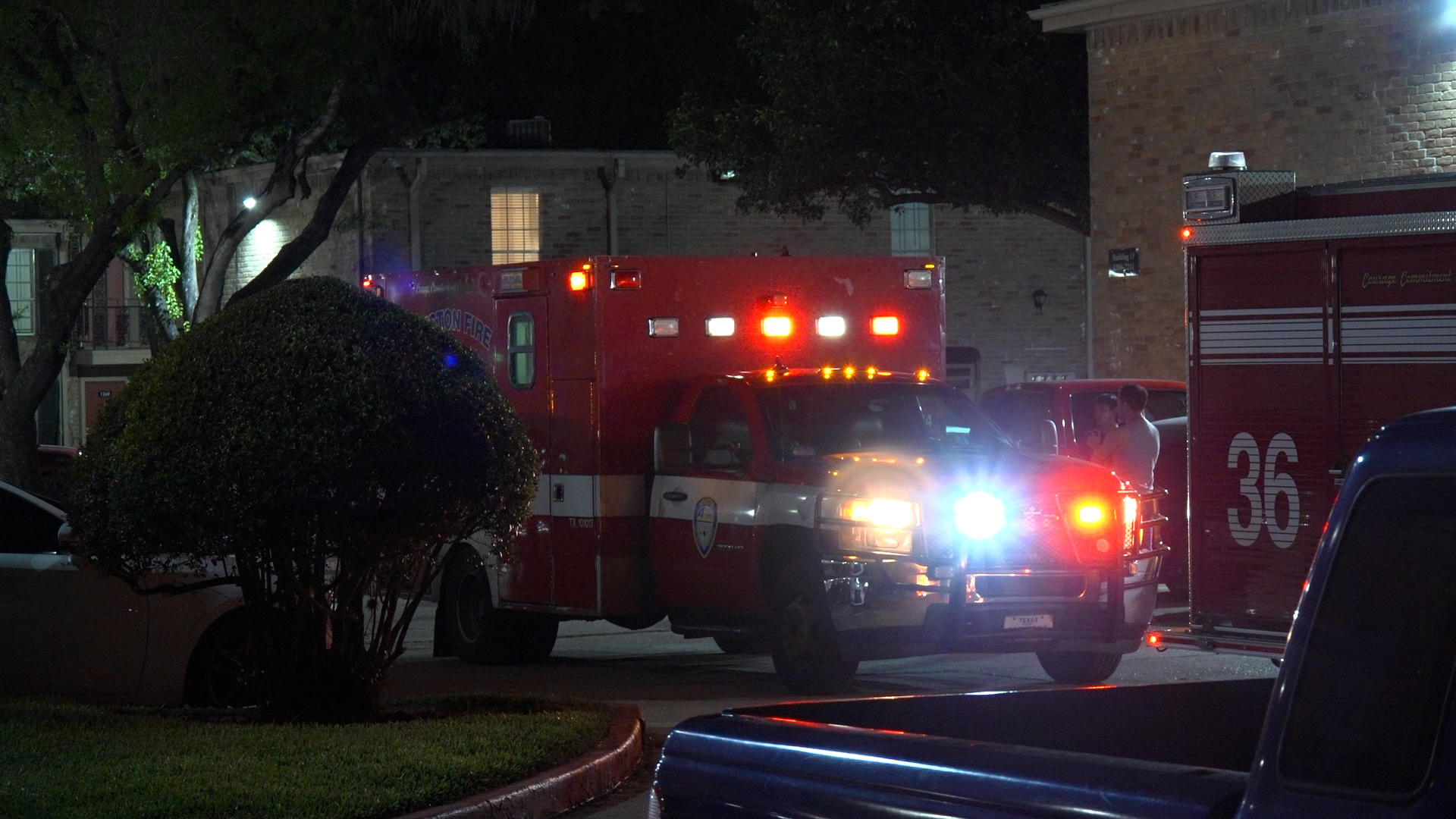 Nighttime emergency scene with a red fire engine and a vehicle in a parking lot, flashing lights visible outside a brick building