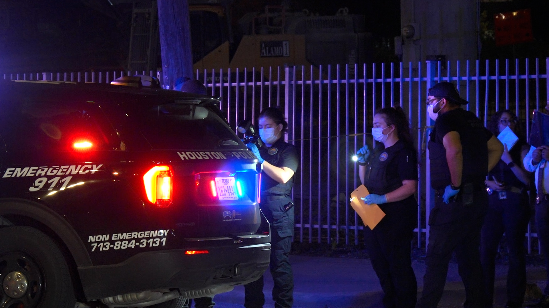 Houston police emergency SUV with red and blue lights at night; masked responders assist a line of people near a fence.