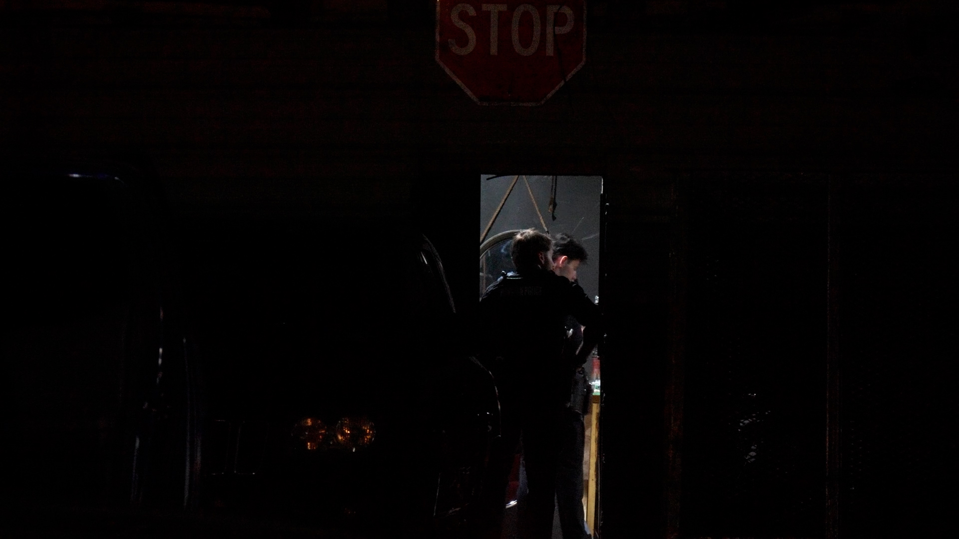Dark night scene with a red STOP sign above a doorway; two people stand inside the lit doorway.