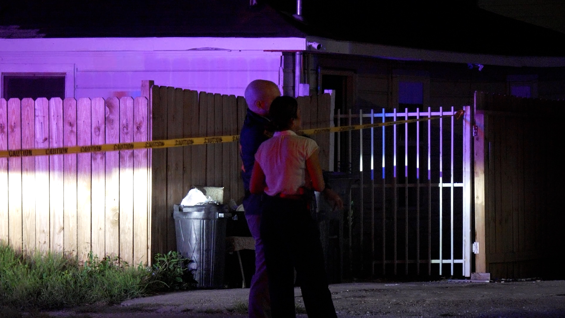 Two people stand by a wooden fence at night, illuminated by purple police lights, with yellow caution tape marking a crime scene behind them.