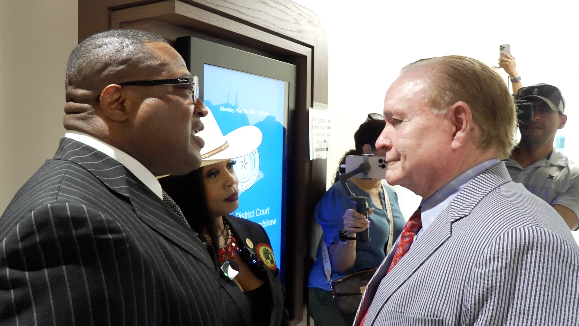 Two men in pinstripe suits face each other in a hallway, with a large blue display screen and people taking photos in the background.
