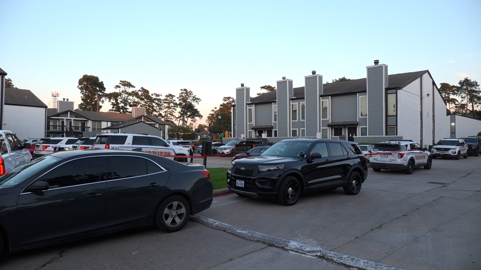 Parking lot in front of a grey apartment complex, with several police and sheriff vehicles and red caution tape across the area.