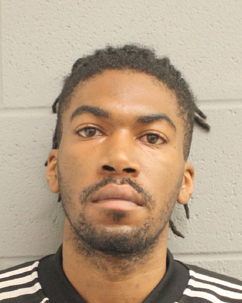 Mugshot of a young man with short dreadlocks and striped clothing against a gray wall, facing camera.