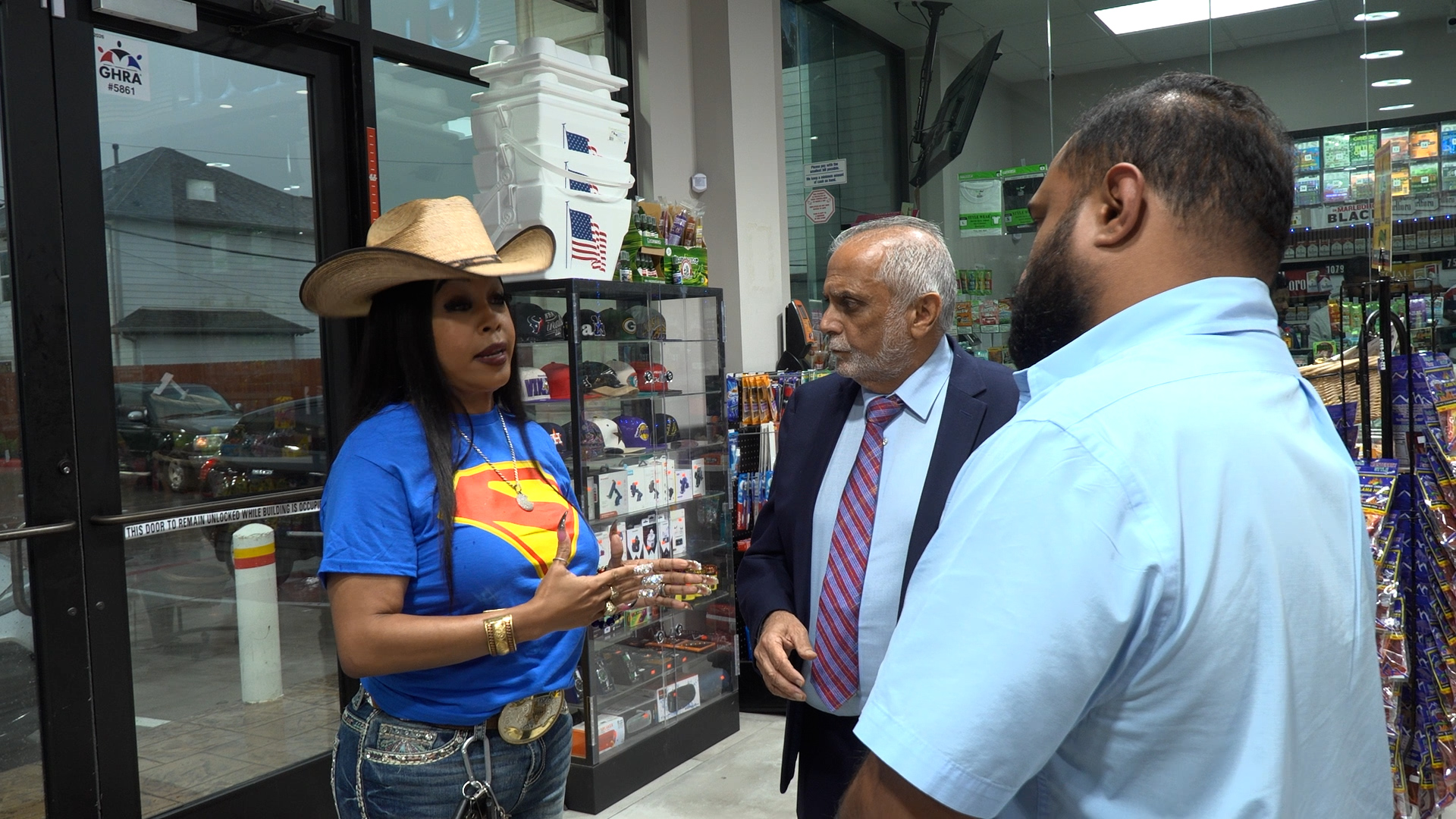 Woman in a cowboy hat and blue Superman shirt speaks to two men inside a convenience store near a display case of merchandise.