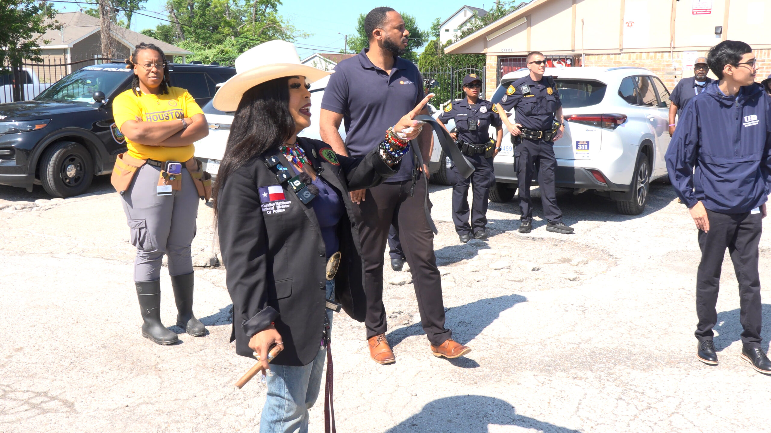 Diverse group of officials and officers in a parking lot; a woman in a black blazer and white hat gestures and speaks while others listen nearby.
