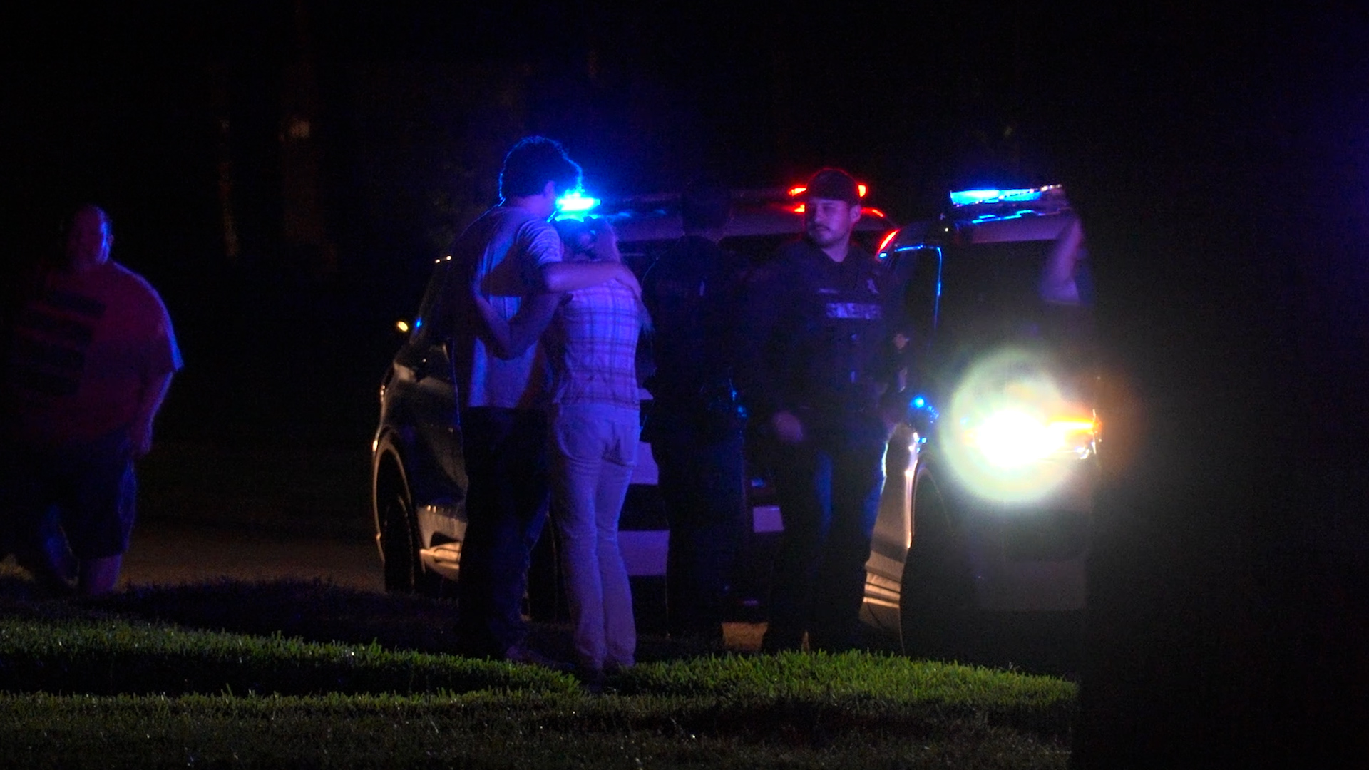 Nighttime scene with a police car and flashing blue/red lights; several people stand nearby, some embracing.