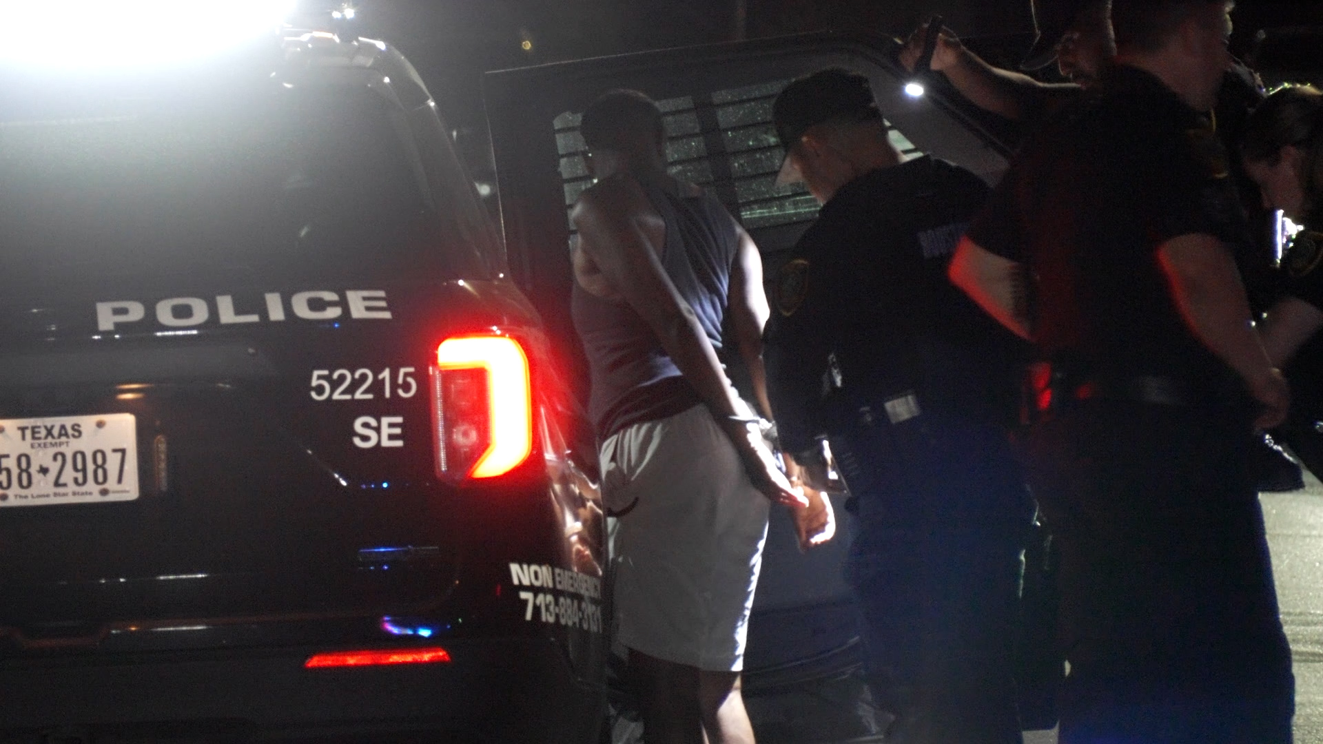 Nighttime scene of a dark police SUV with 'POLICE' on the side, a shirtless man with hands behind his back being detained by officers nearby, flashing lights illuminating the scene