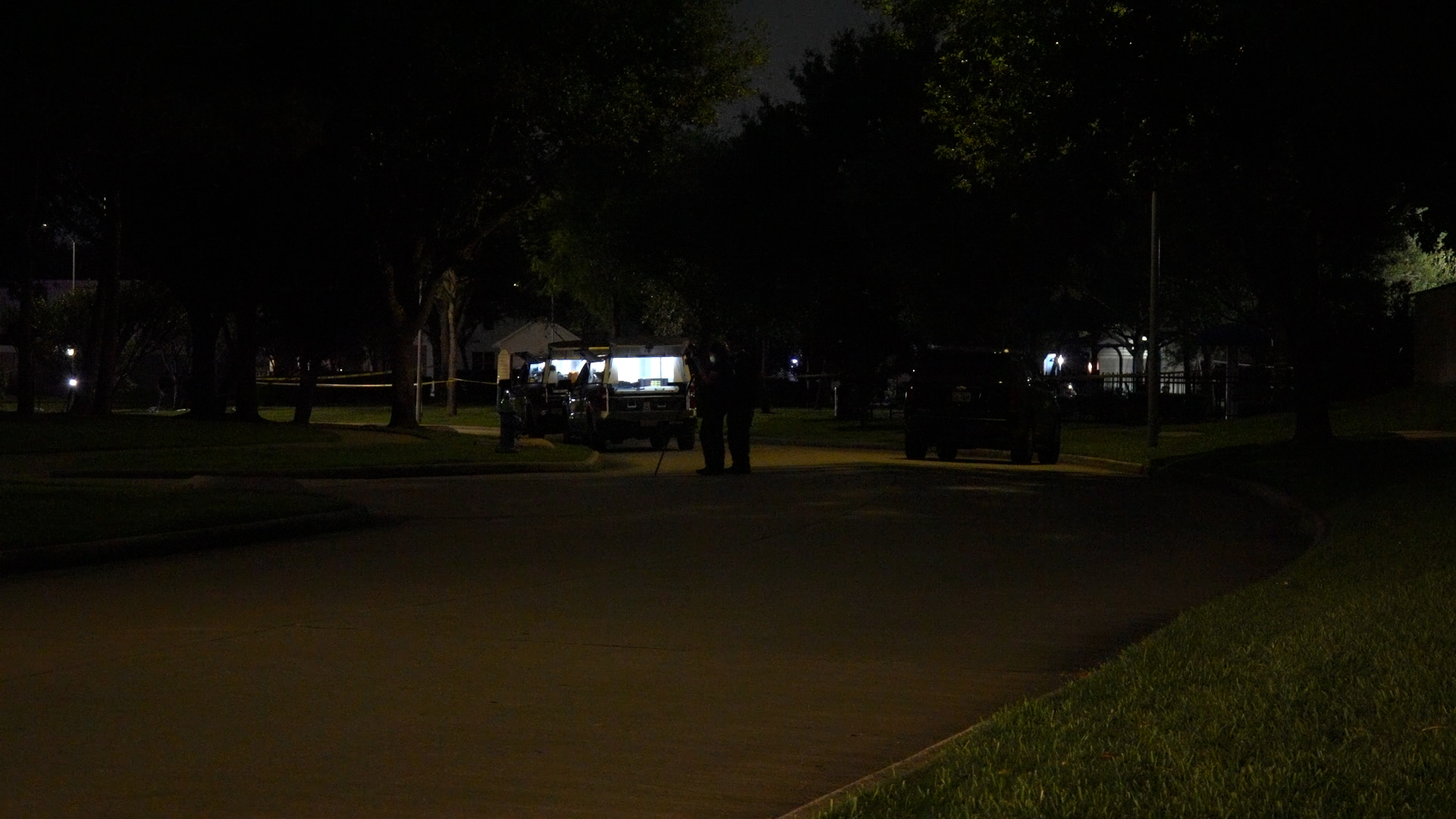 Night scene of a residential street with police SUVs and a lit tented command post set up under trees at night