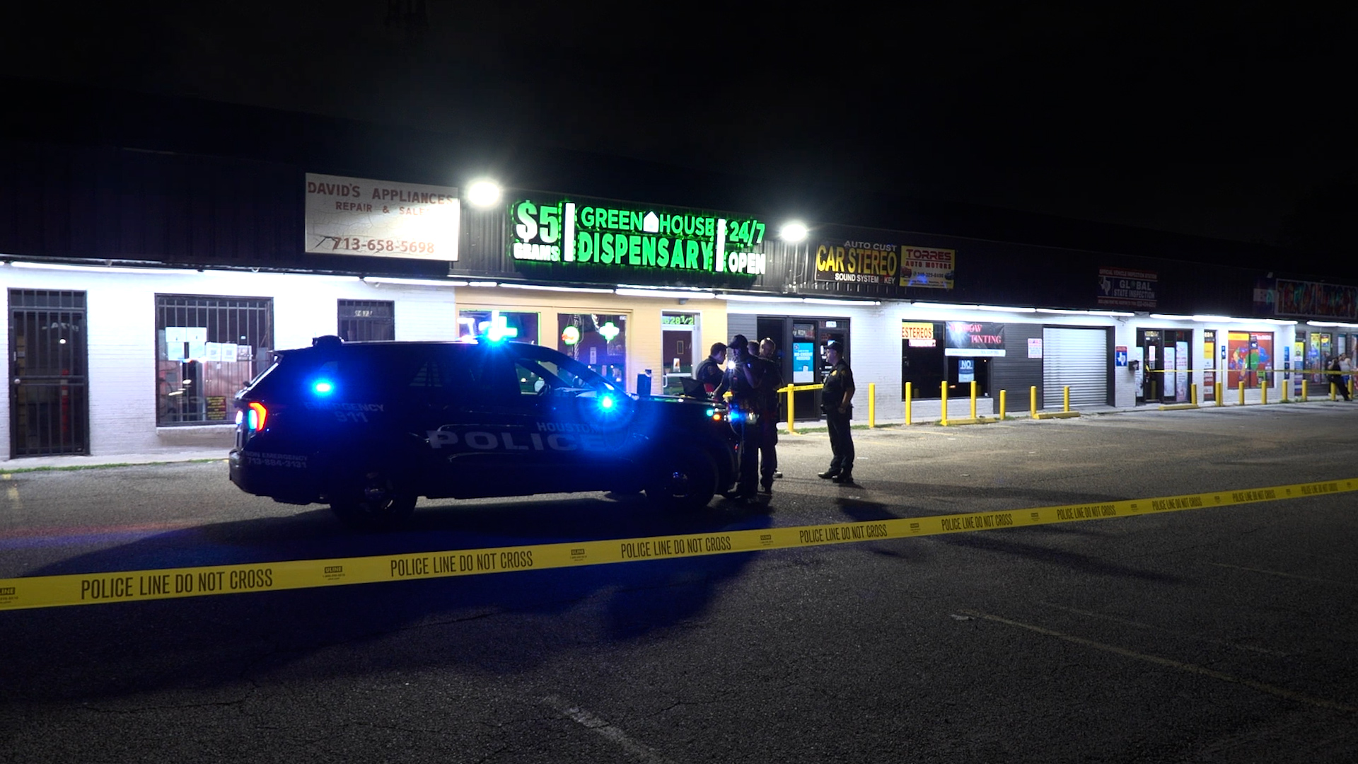 Nighttime scene outside a strip mall with a police SUV and flashing blue lights, taped off by yellow 'POLICE LINE DO NOT CROSS' lines and officers present outside storefronts.