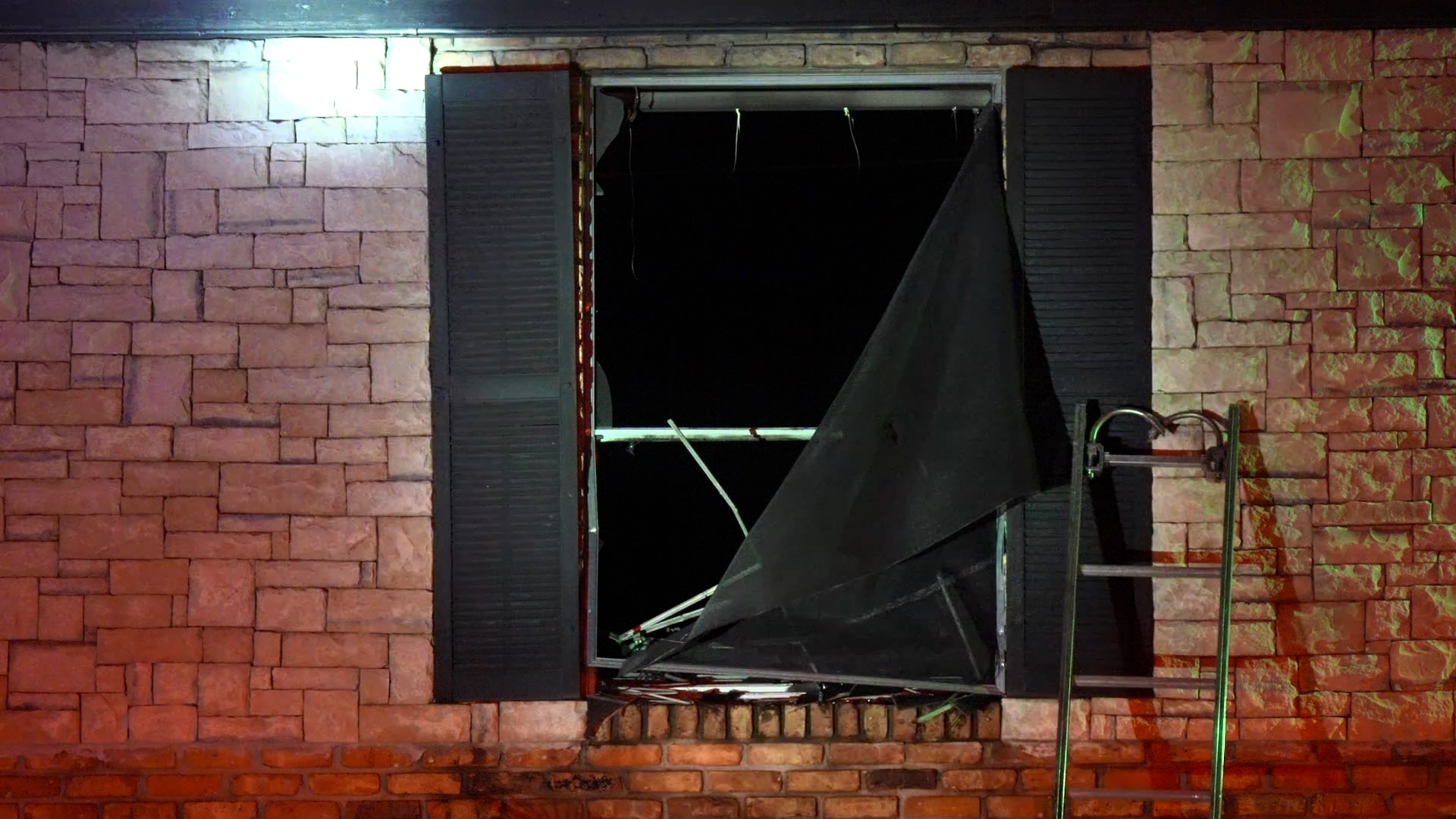 Broken window in a brick exterior wall with a torn black tarp draped across the opening and scattered debris on the sill; a ladder and a green hose appear on the right.