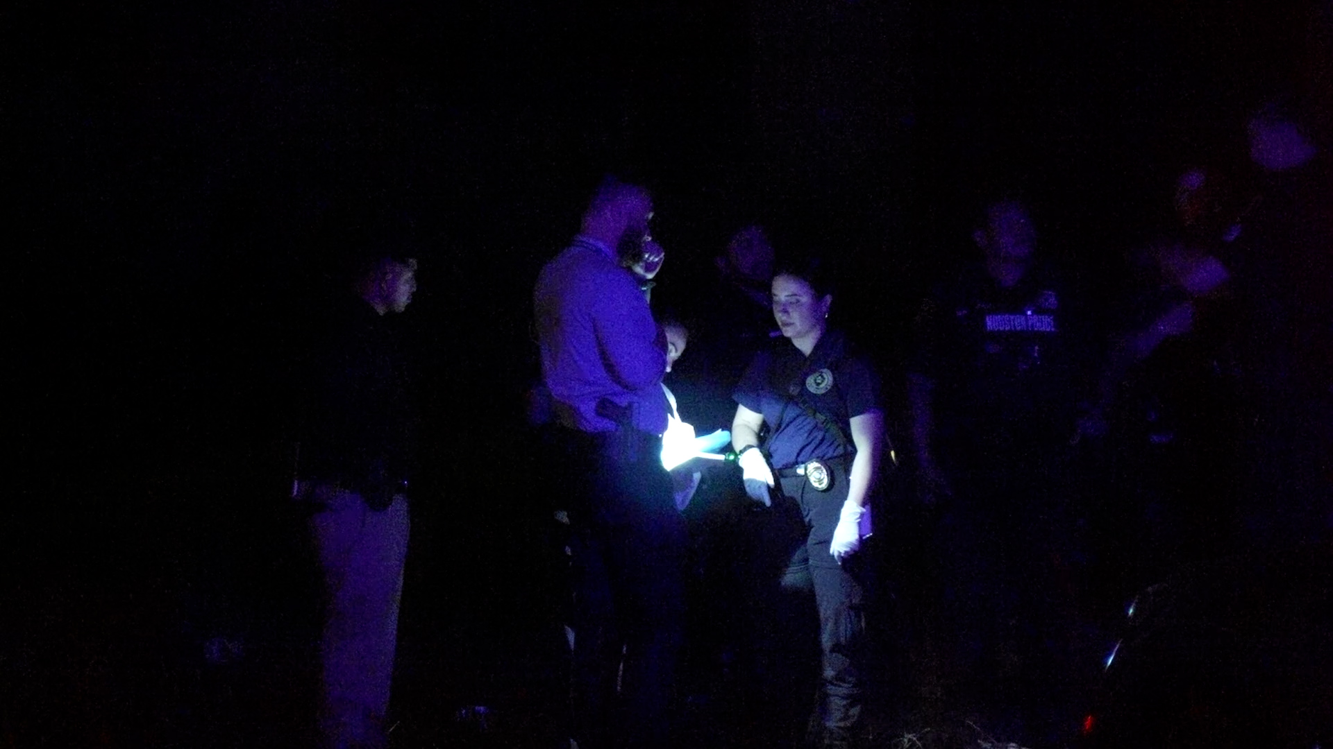 Police officers and onlookers in a dark room, illuminated by a flashlight as they examine something on a table.