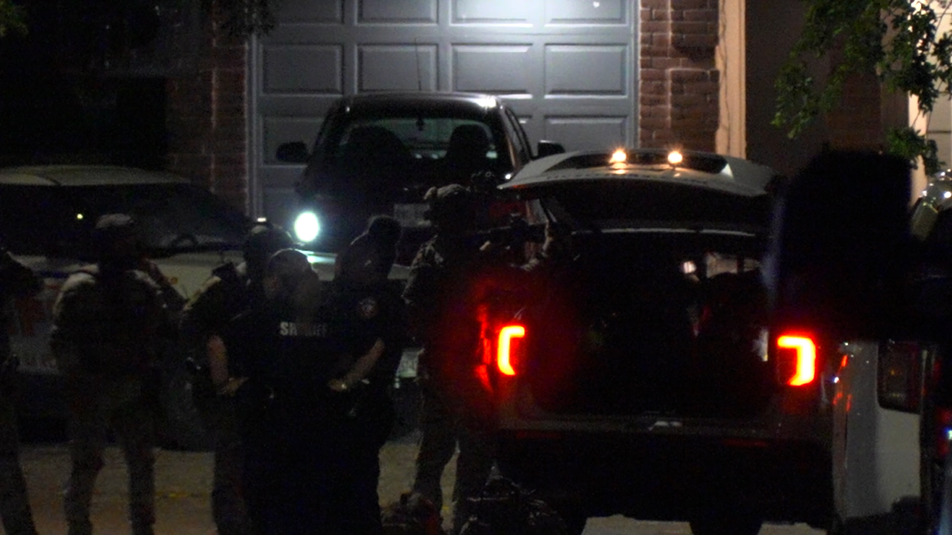 Nighttime scene showing a parked SUV and a larger vehicle with flashing lights, surrounded by armed police in tactical gear outside a residential garage.
