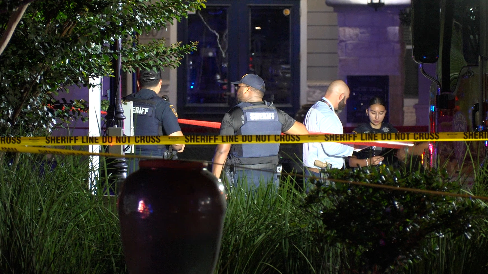 Sheriff deputies stand behind yellow caution tape at a nighttime outdoor scene with greenery in the foreground.