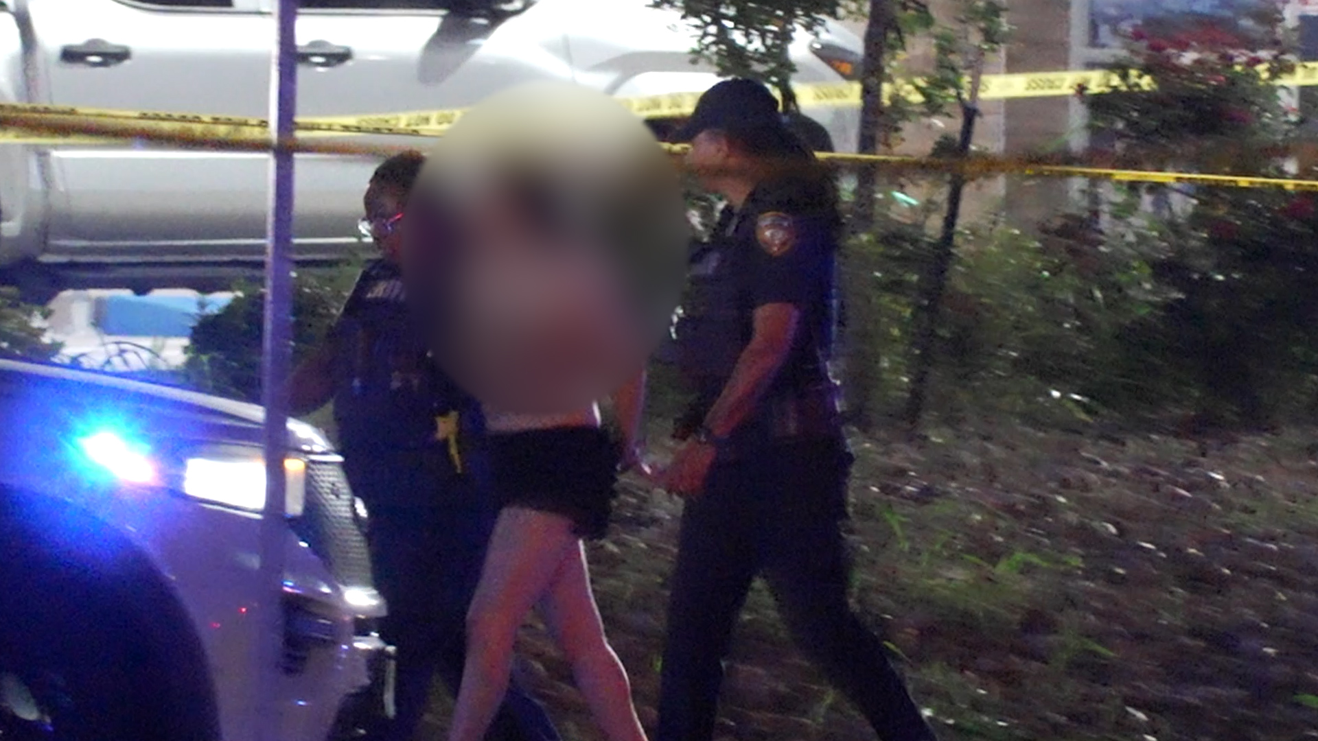 Police officers at a night-time crime scene with yellow caution tape and a vehicle with flashing blue lights in the foreground; a blurred person is being escorted.